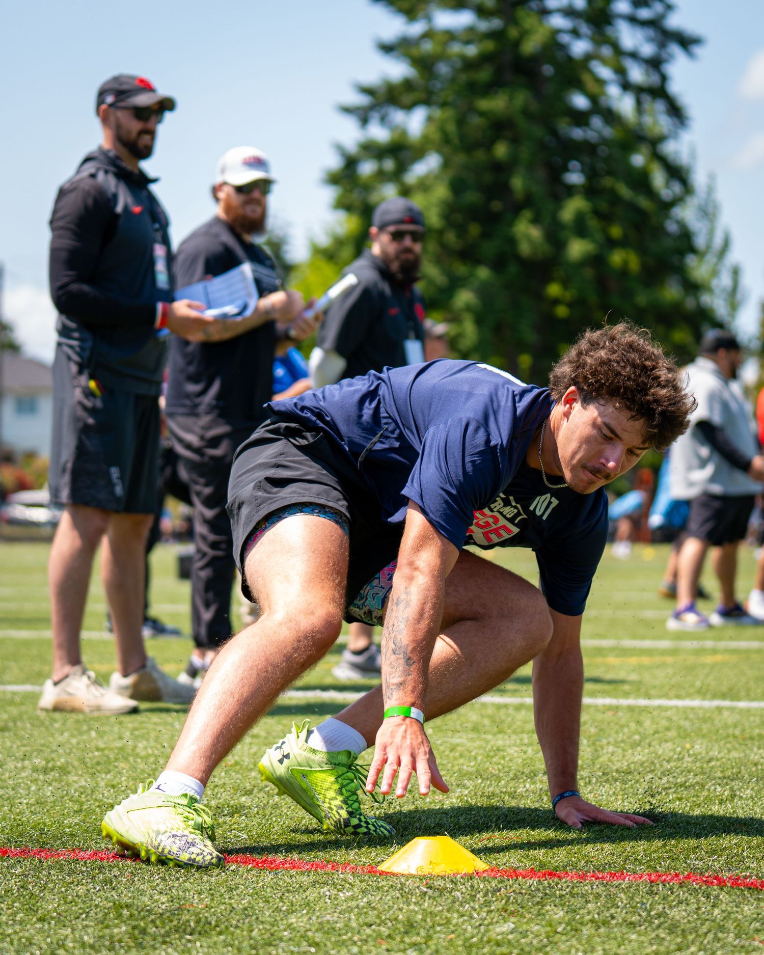 Athlete performs agility drill with yellow cone on green turf field during football combine workout with coaches observing.