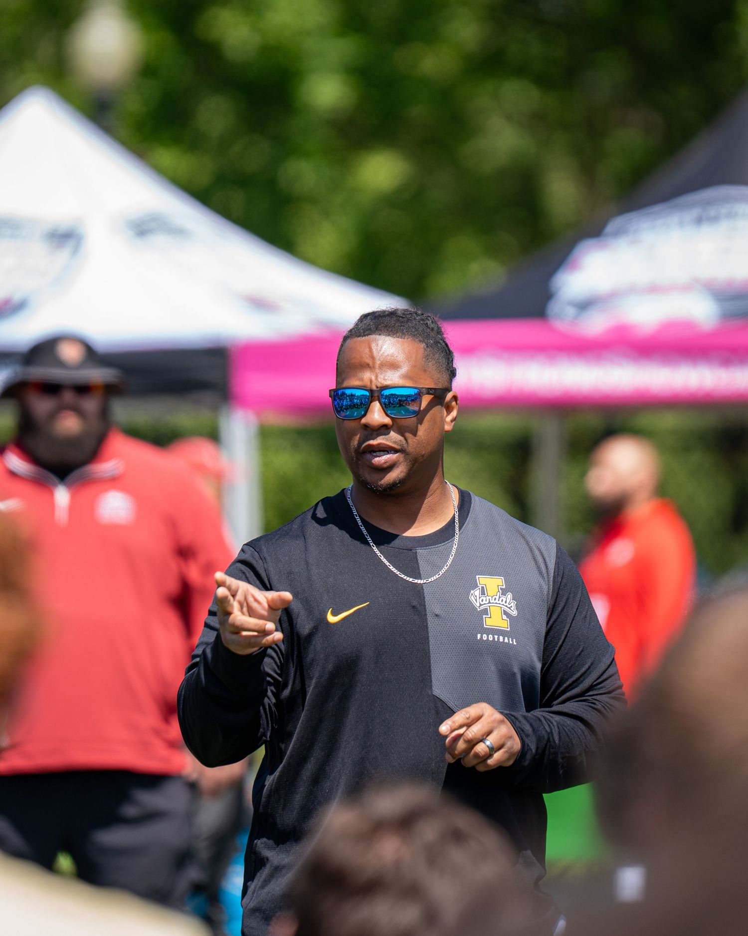 A football coach in a black Nike shirt and sunglasses gestures while speaking outdoors at a sunny event.