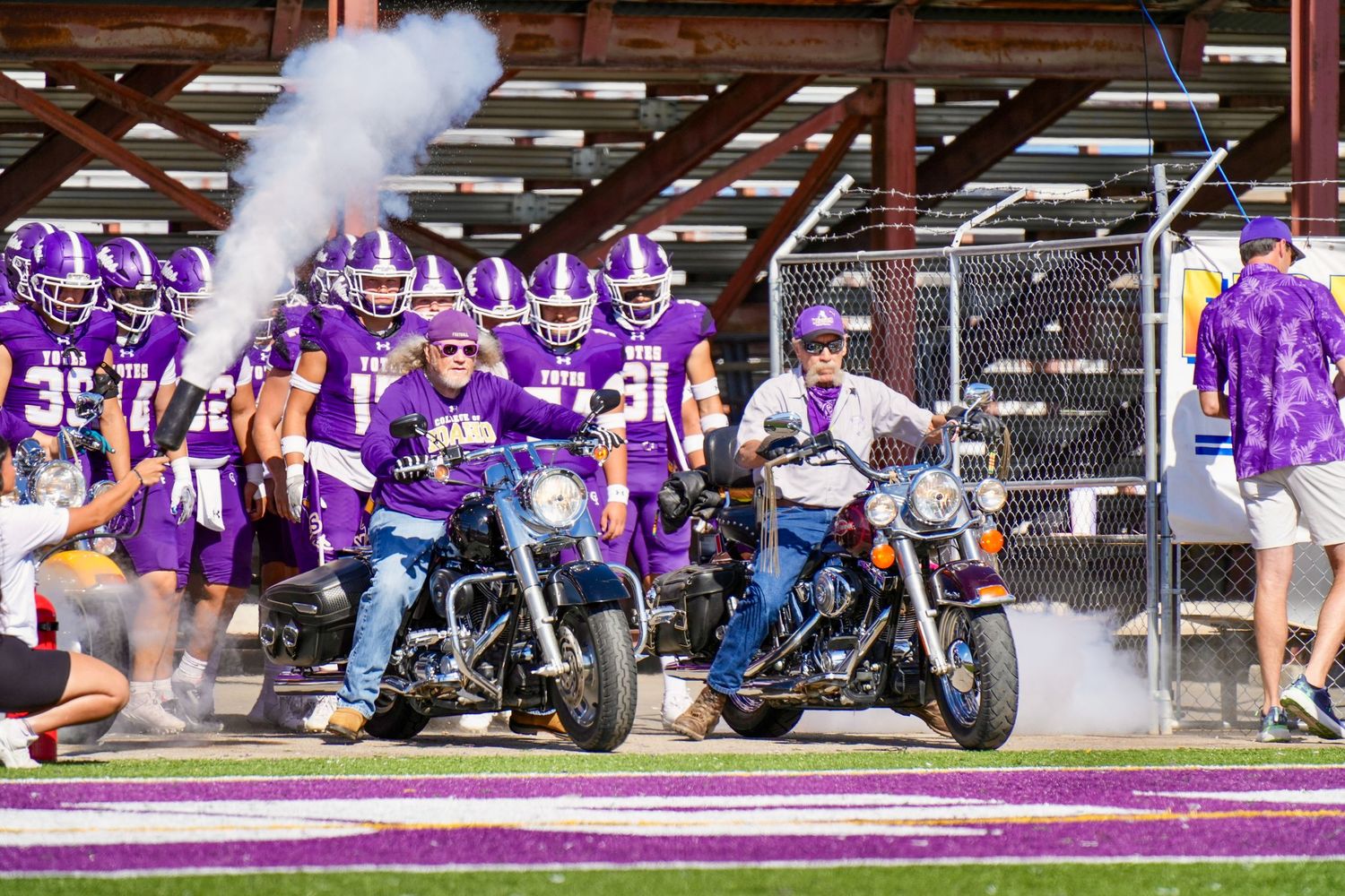 Two motorcycles rev engines creating smoke while purple-clad fans cheer from bleachers at a sports event.