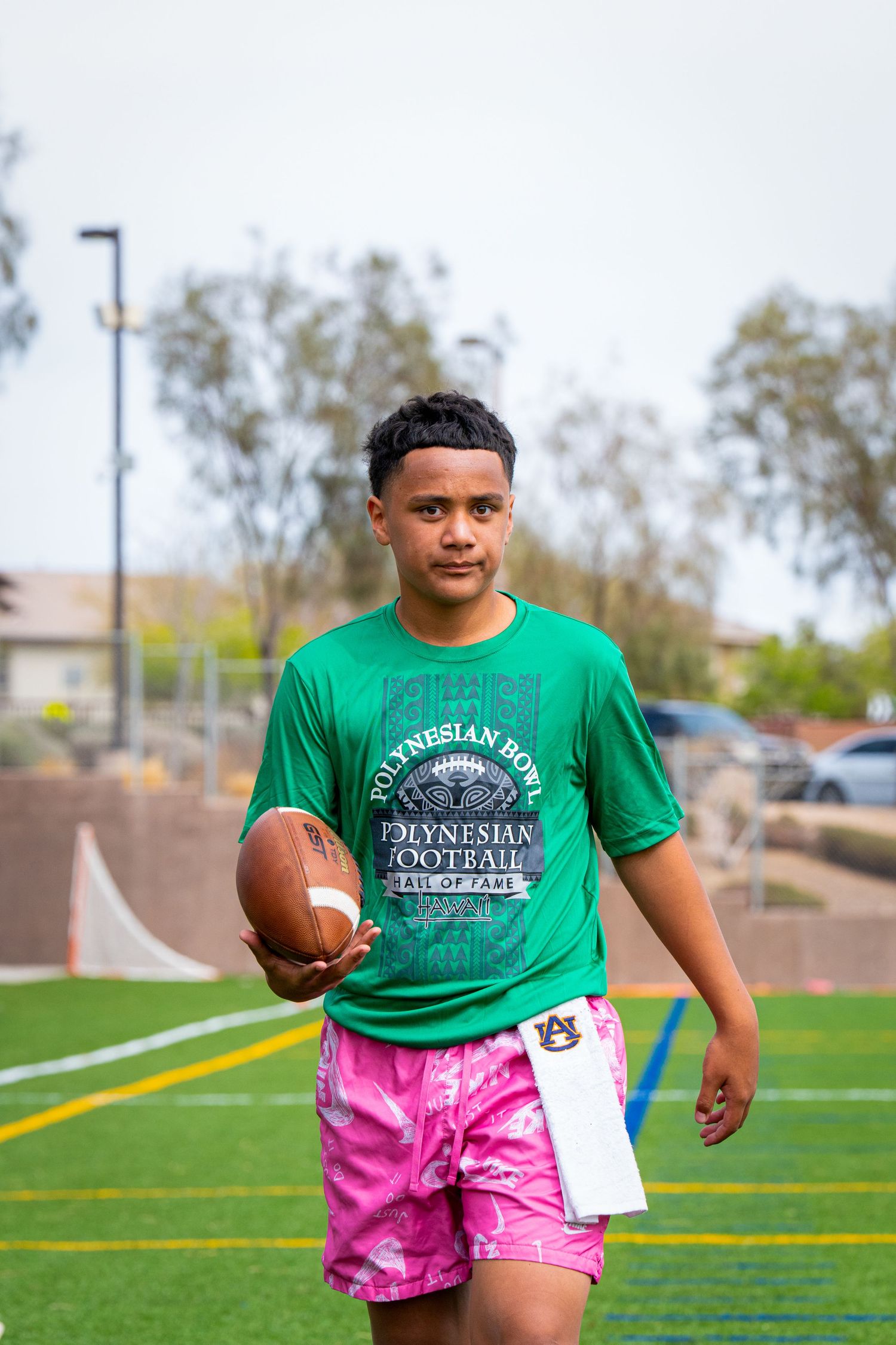 Athlete poses with football wearing green Polynesian Football shirt and pink shorts on field.
