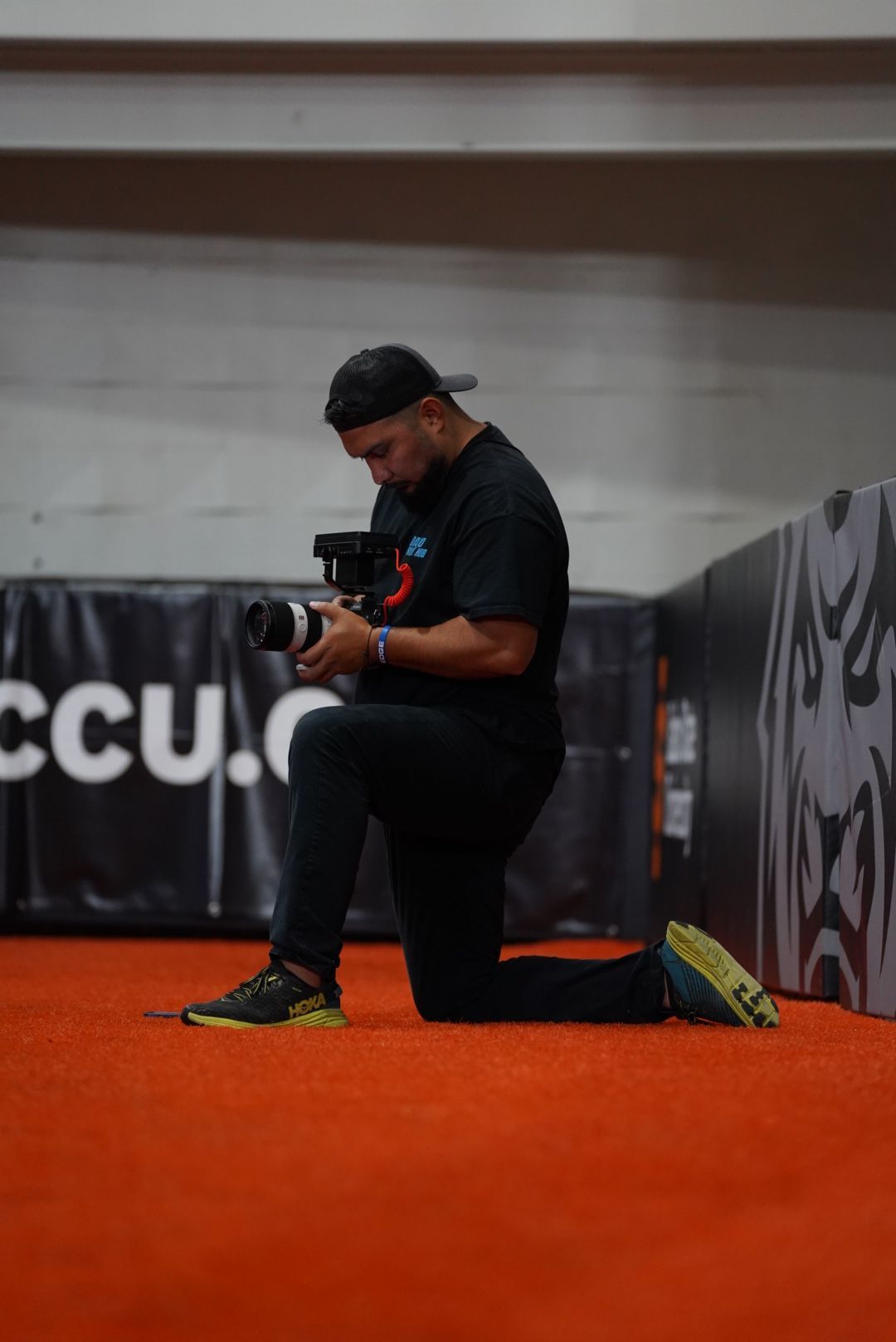 Photographer in black attire crouches on bright orange floor while adjusting camera settings in dimly lit studio.