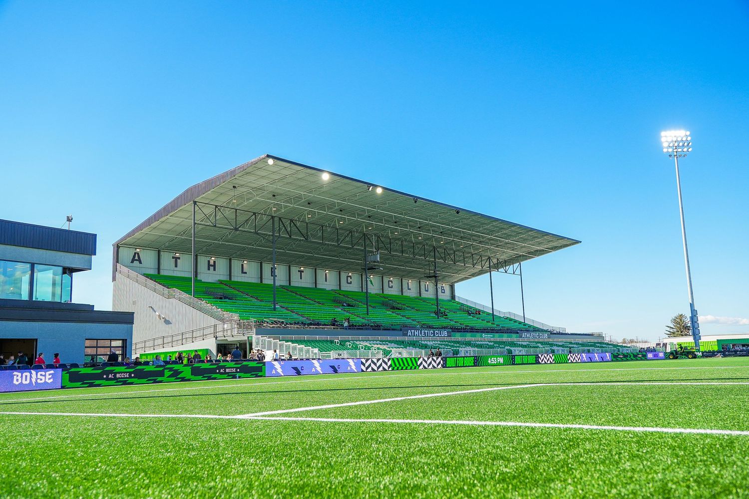 Modern football stadium with green pitch, covered grandstand, and bright floodlights under a clear blue sky.