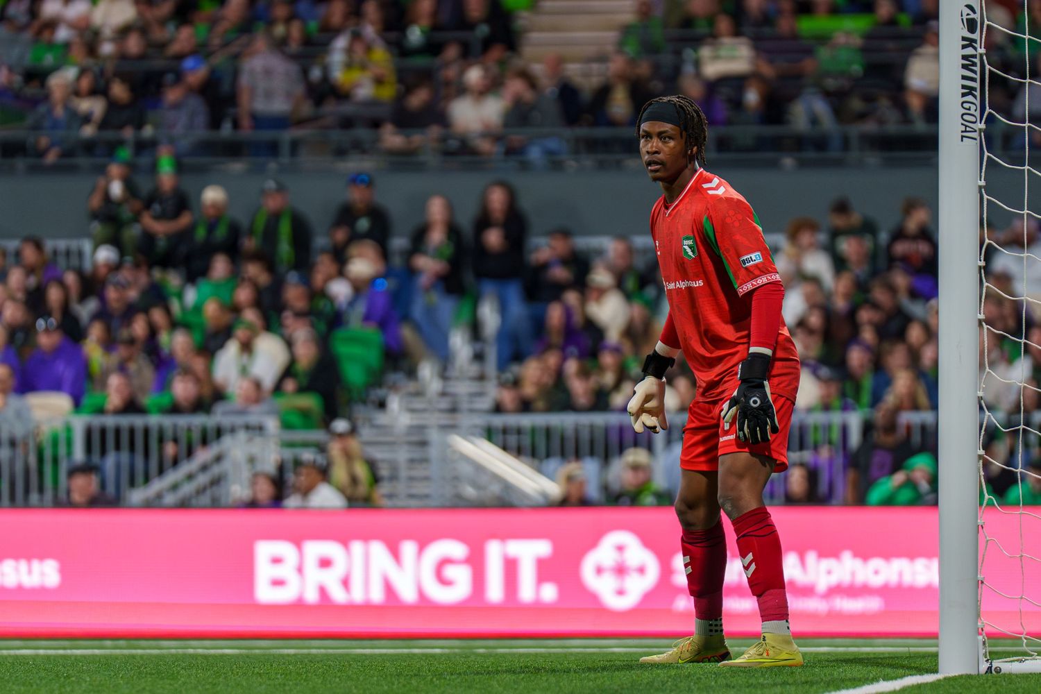 Soccer goalkeeper in red uniform stands alert near the goal post during a professional MLS match at a green stadium.