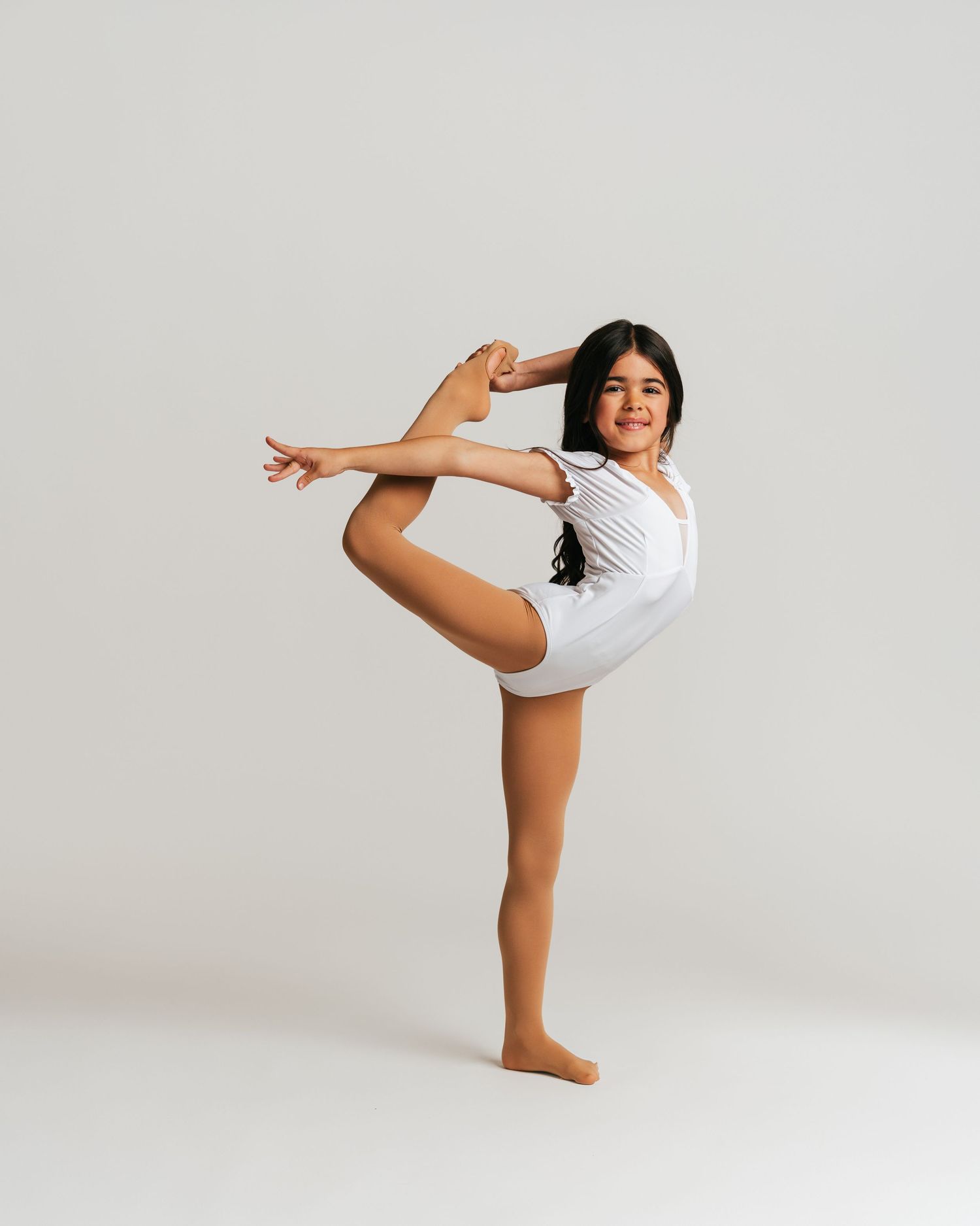 Young dancer in white leotard smiling while doing a standing split hold in studio by Bylitos Studio Michigan