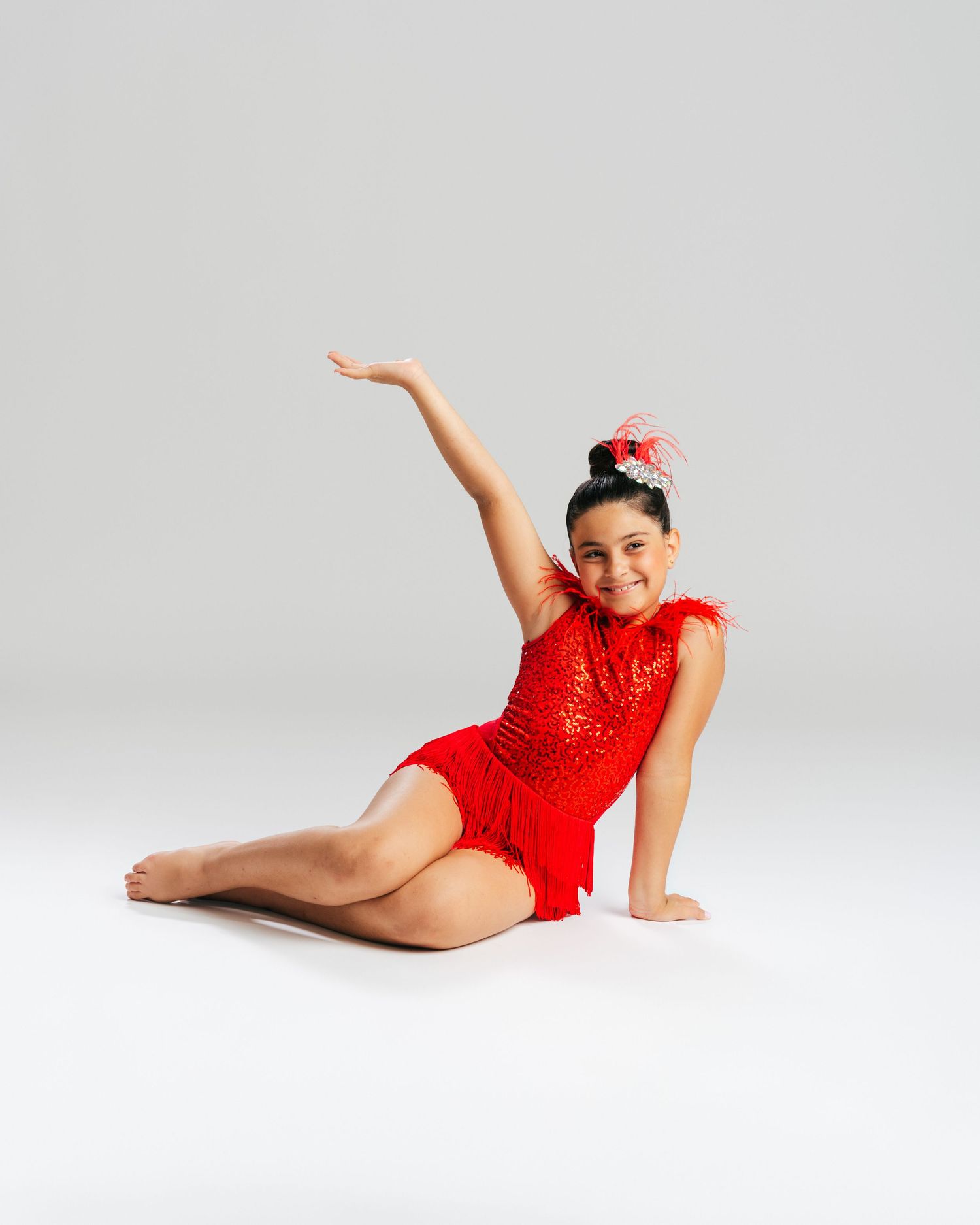 Young dancer in red sequin costume seated on floor with arm raised during studio editorial by Bylitos Studio Michigan