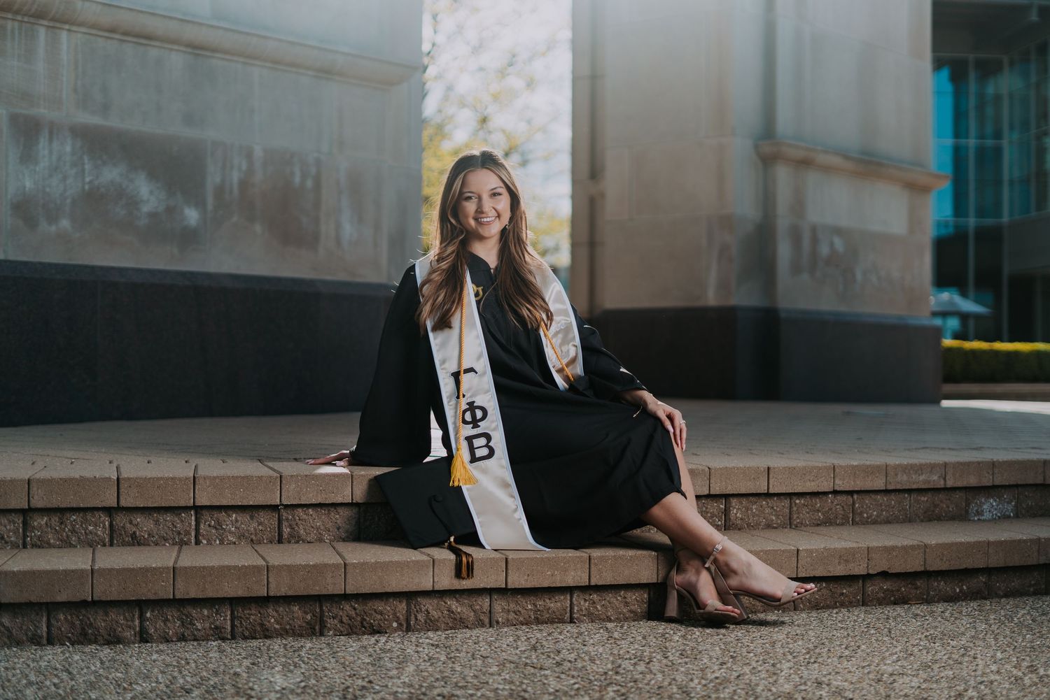 Graduating senior in cap and gown with sorority sash seated on steps by Bylitos Studio Michigan