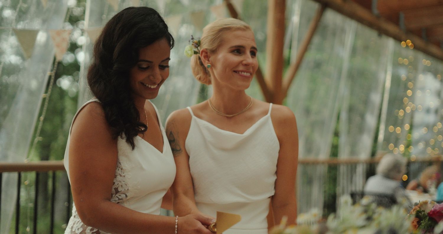 Two brides smiling together at wedding reception under string lights by Bylitos Studio Michigan