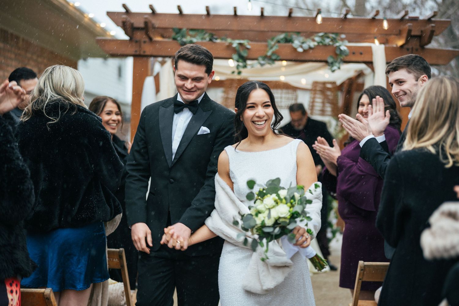 Newlyweds walking down the aisle as guests cheer under a wooden pergola by Bylitos Studio Michigan