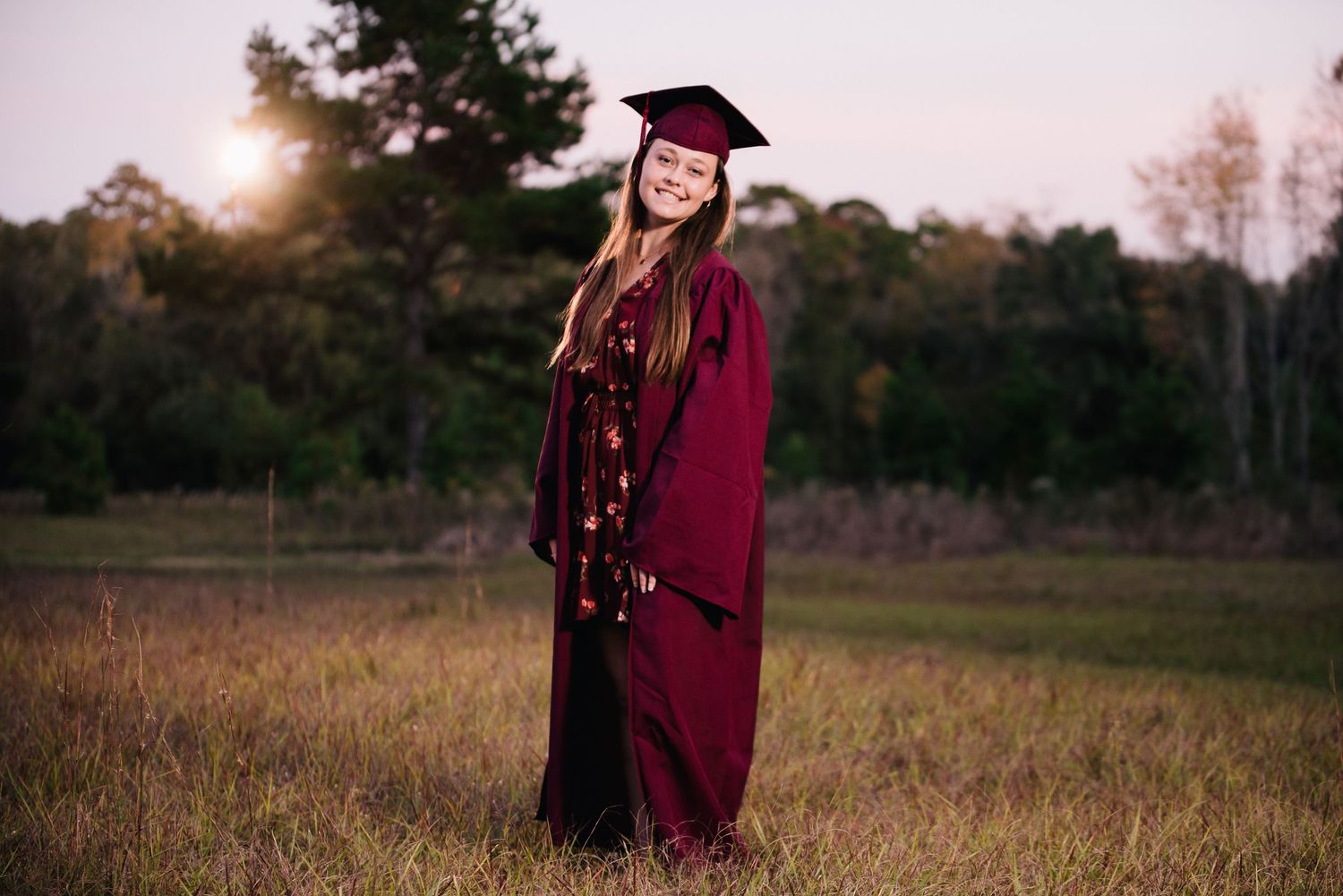 Graduate poses in maroon academic regalia against a scenic autumn field at sunset.