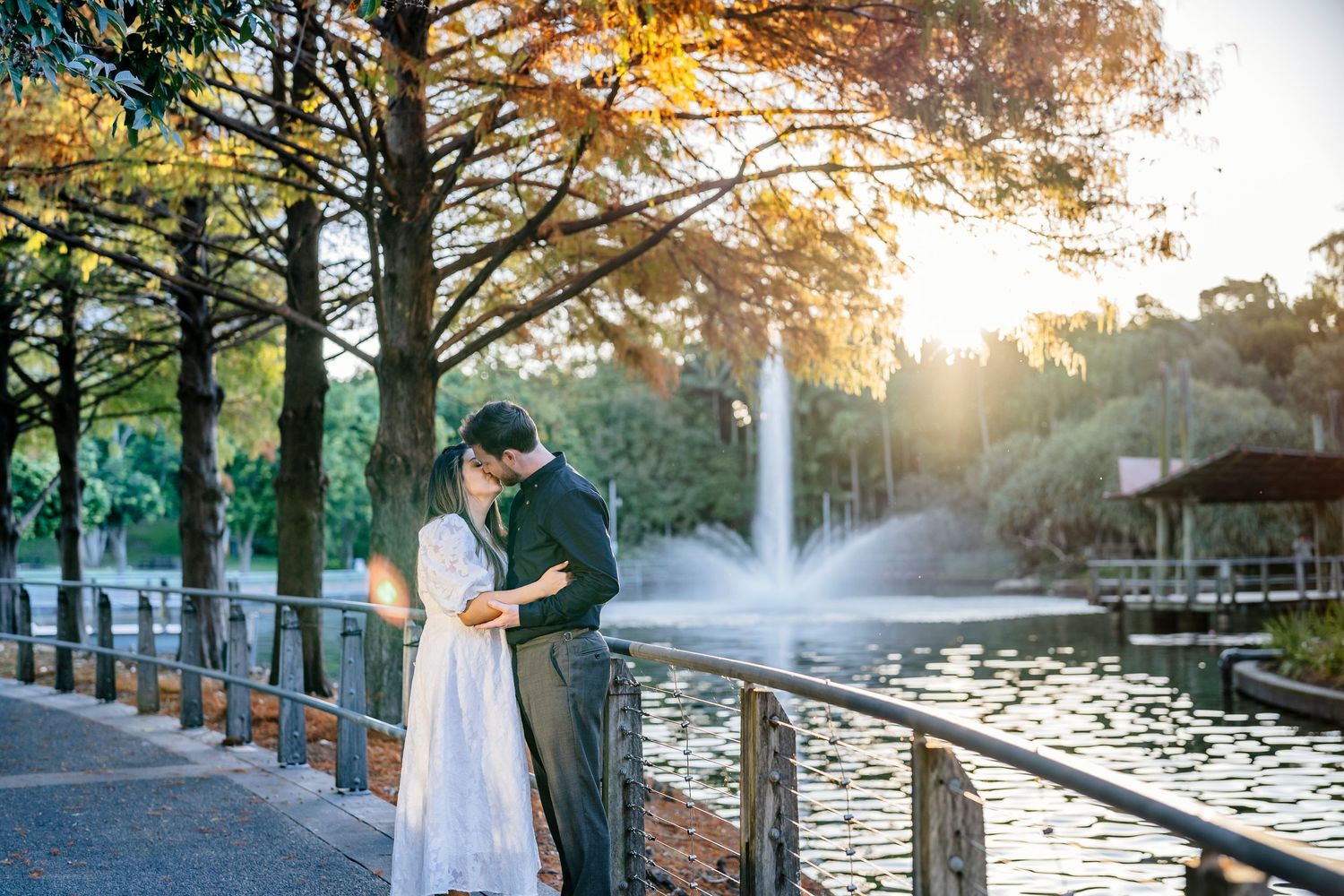 Couple sharing a romantic moment by a serene pond with a fountain, surrounded by autumn trees at golden hour.