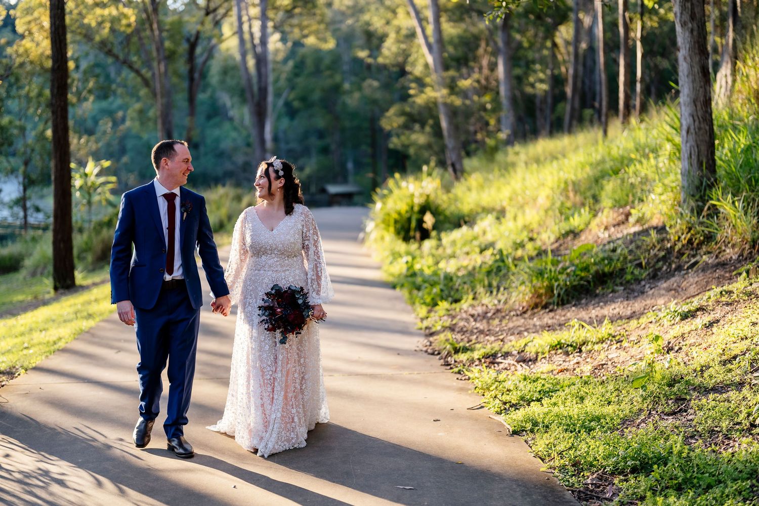 Bride and groom walking hand in hand along a sunlit path surrounded by lush greenery on their wedding day at Walkabout Creek in Brisbane