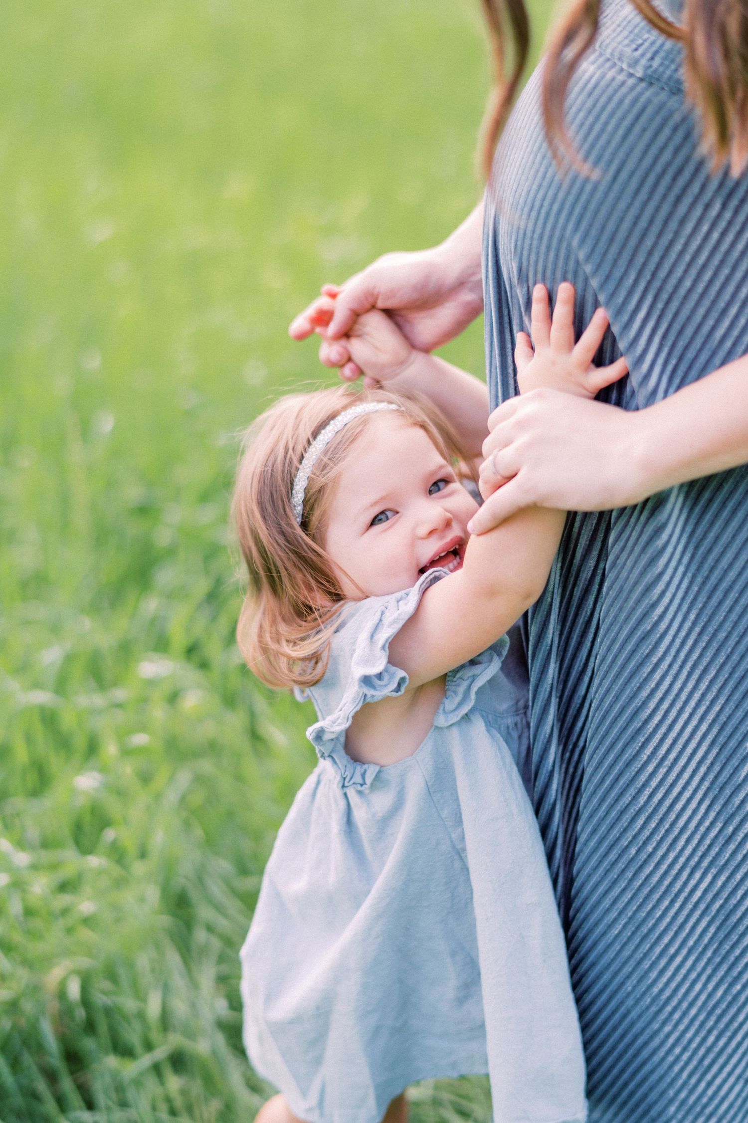 A small child in a blue dress reaches up playfully towards someone in a striped dress while standing in green grass.