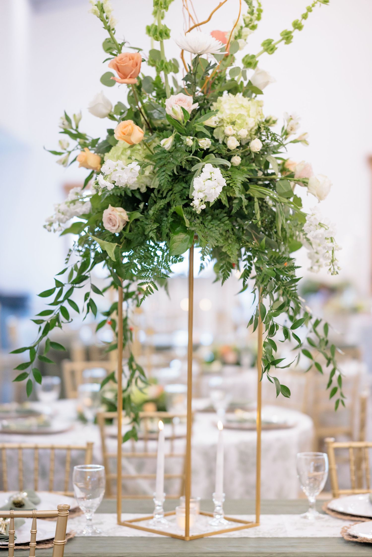 Elegant wedding centerpiece with peach roses and white hydrangeas on tall gold geometric stand with cascading greenery.