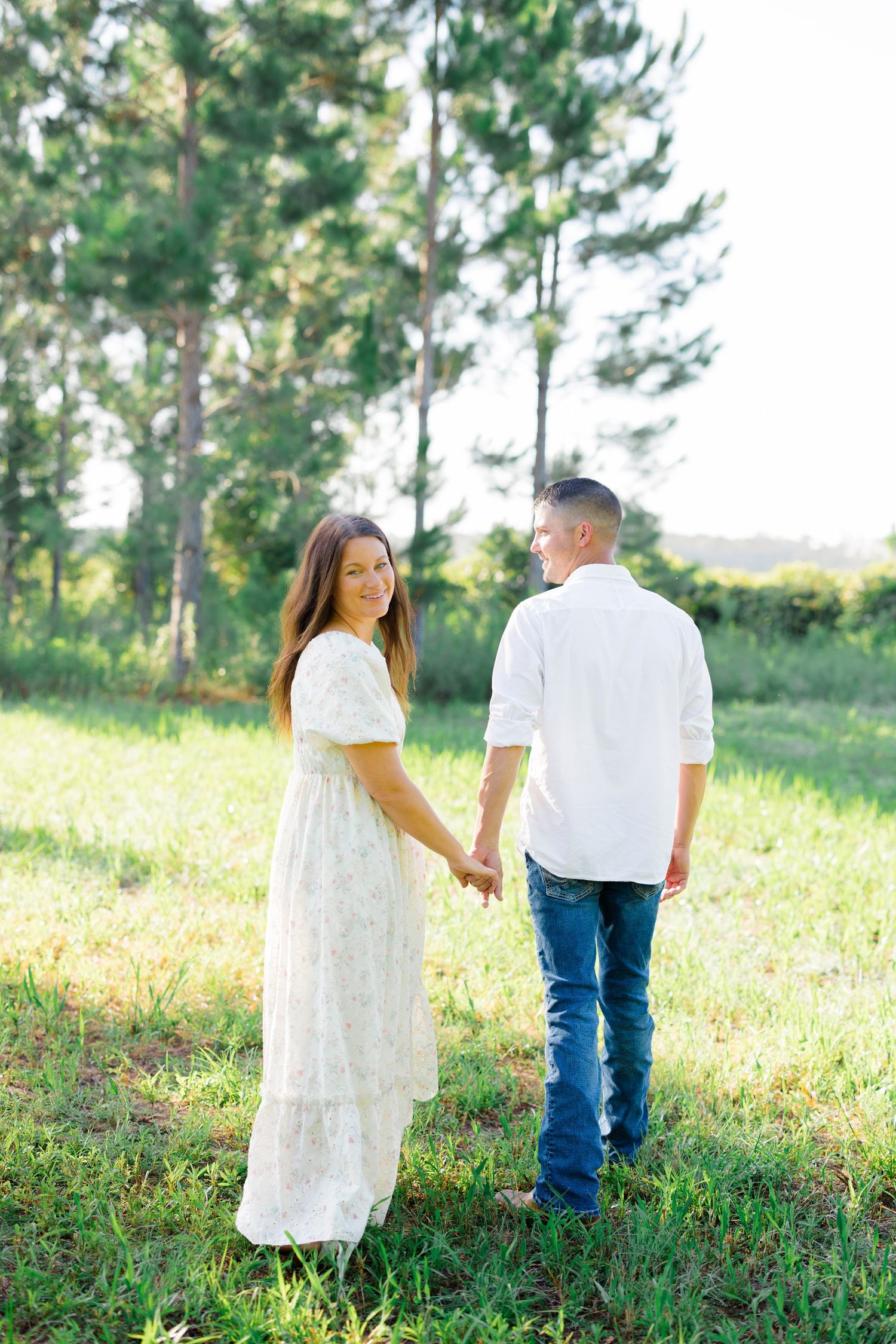 Couple holding hands in a grassy field with pine trees in the background.