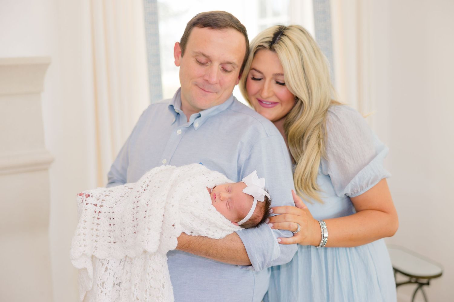 A couple in light blue and white outfits share tender moments together while holding a small baby.