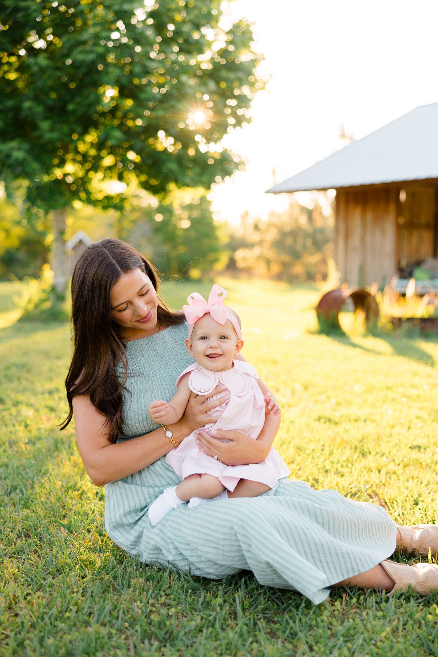 A tender moment between a woman in mint dress and baby in pink outfit sitting on sunny grass near a rustic barn.