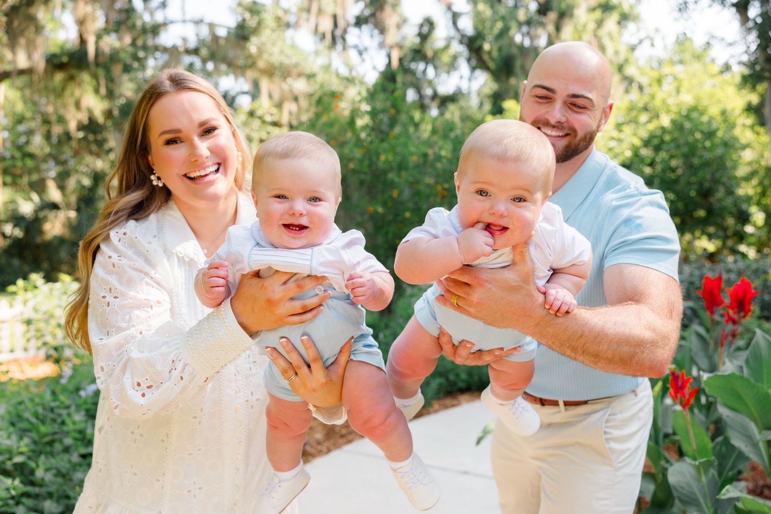 A family holds two smiling babies outdoors in a garden setting with lush greenery in the background.