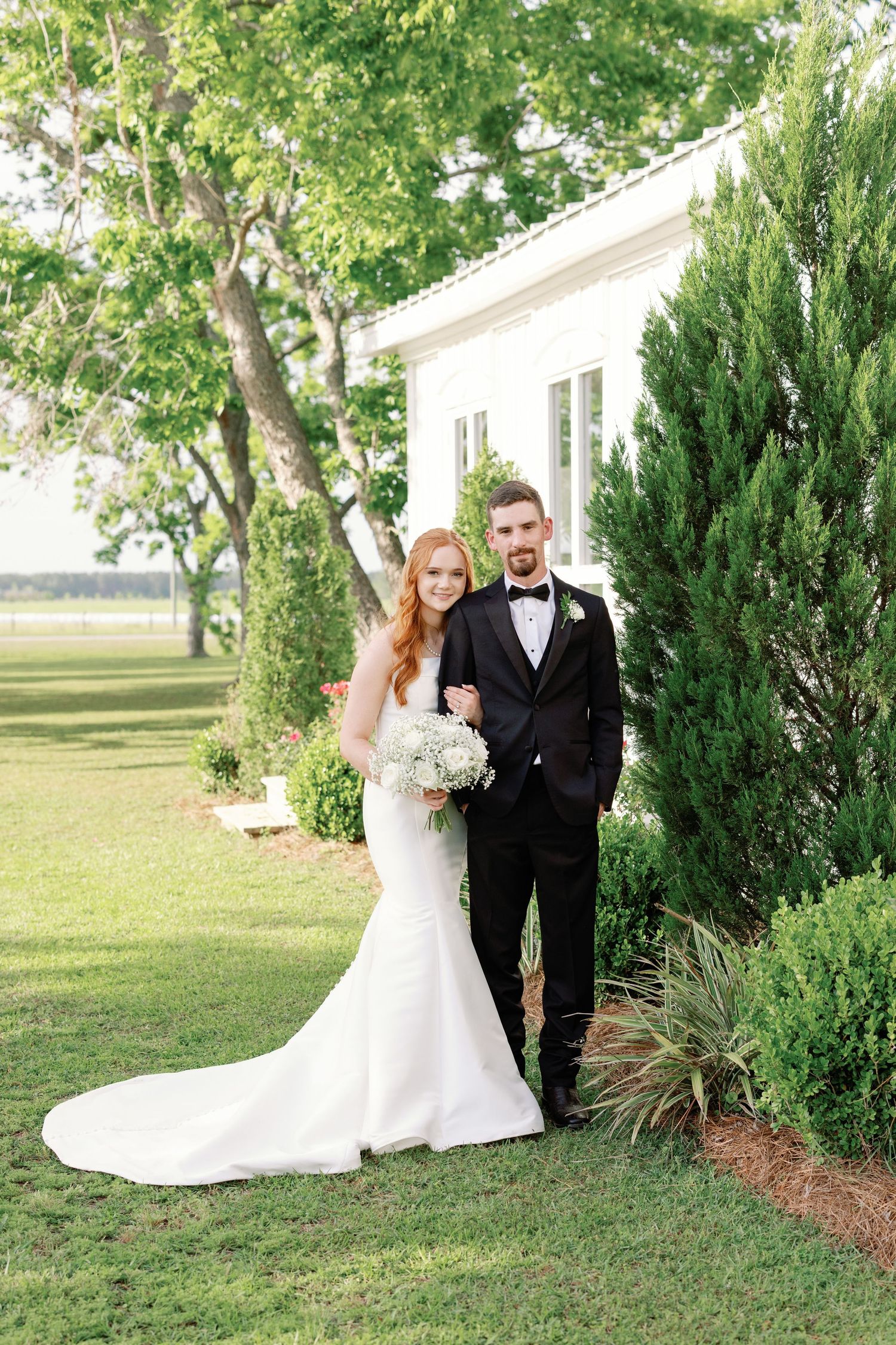 Elegant wedding couple poses outdoors by evergreen bushes in formal attire with flowing white dress.