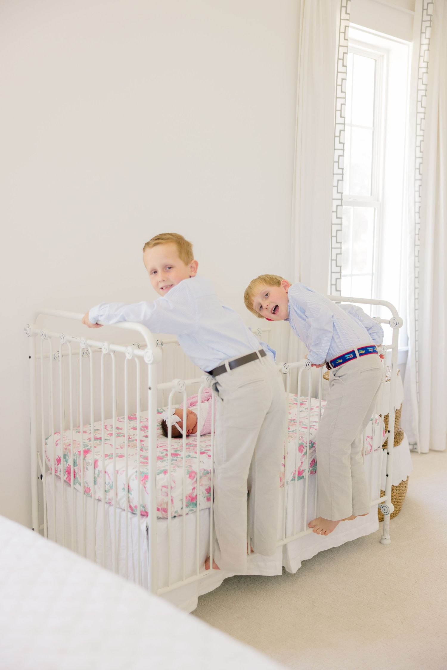 A young child in white pajamas plays and climbs on a white crib with floral bedding in a bright room.