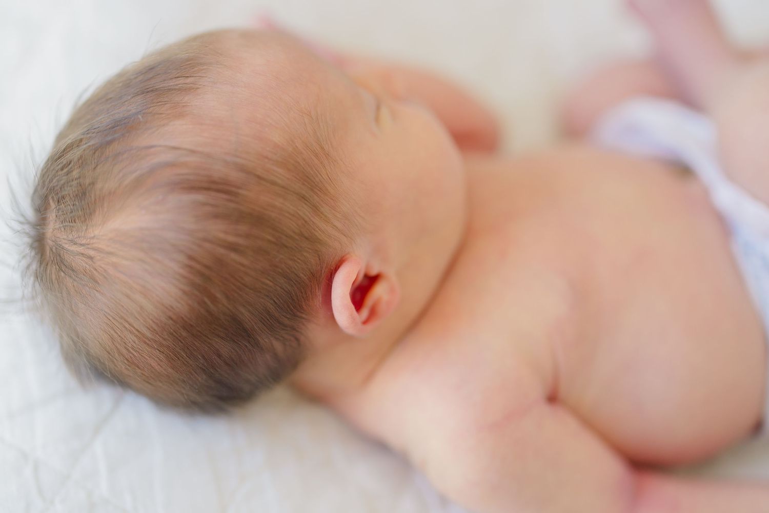 A sleeping newborn baby lies on white bedding, showing their tiny ear and soft brown hair.