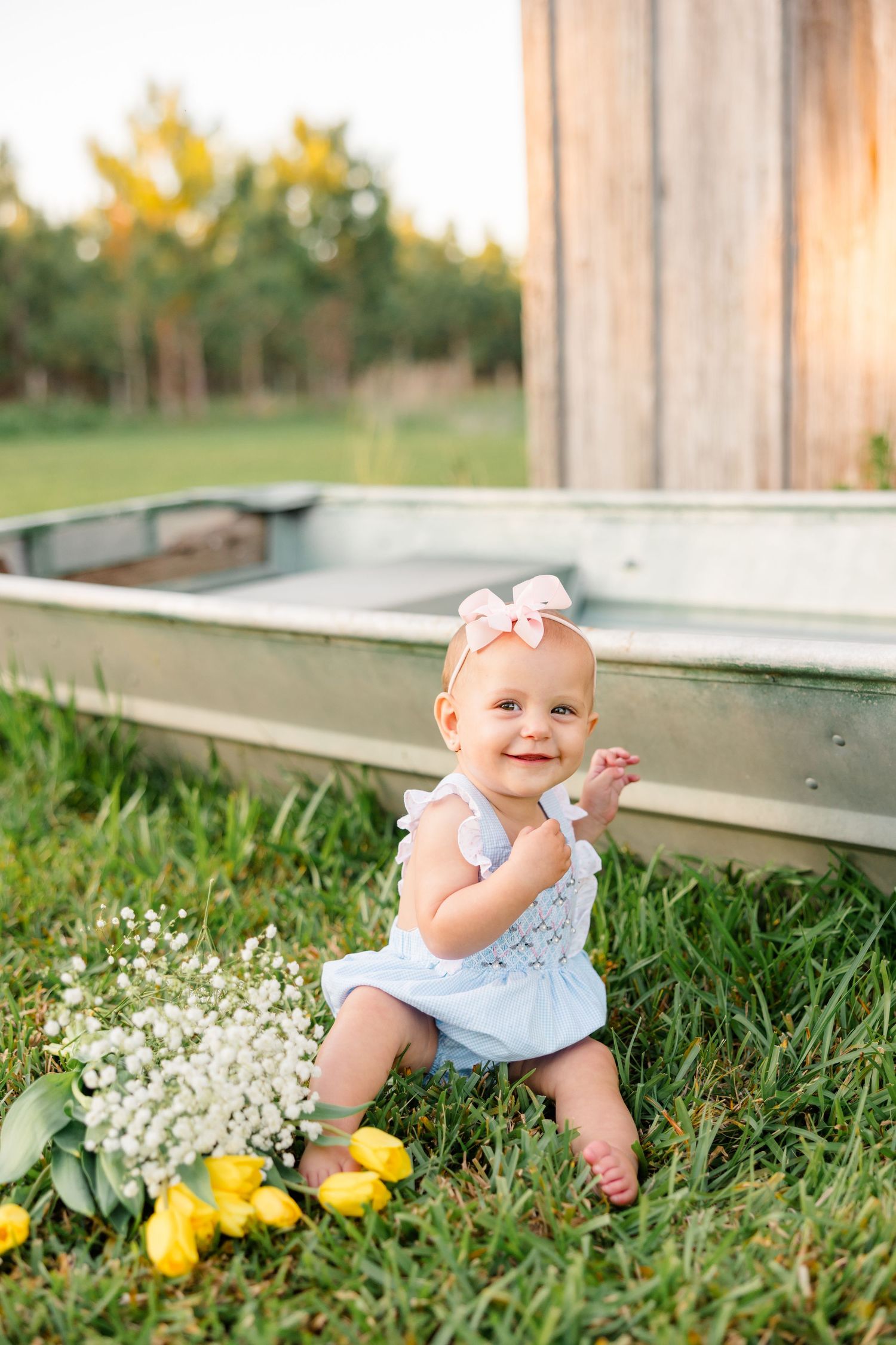 A baby in a blue outfit and pink bow sits on grass next to flowers and citrus fruits.