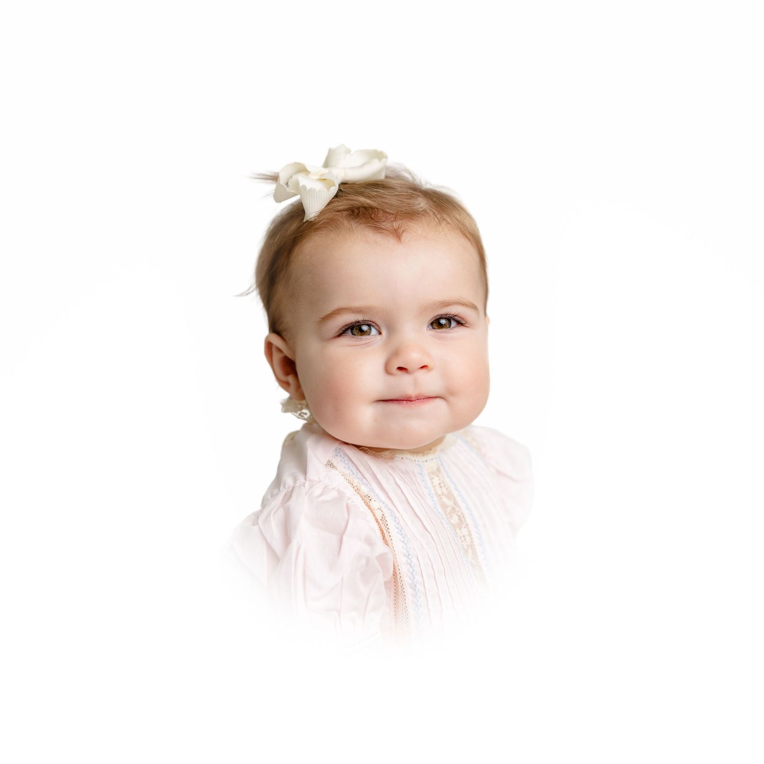 Baby wearing white dress and small bow hair accessory smiles sweetly against white background.