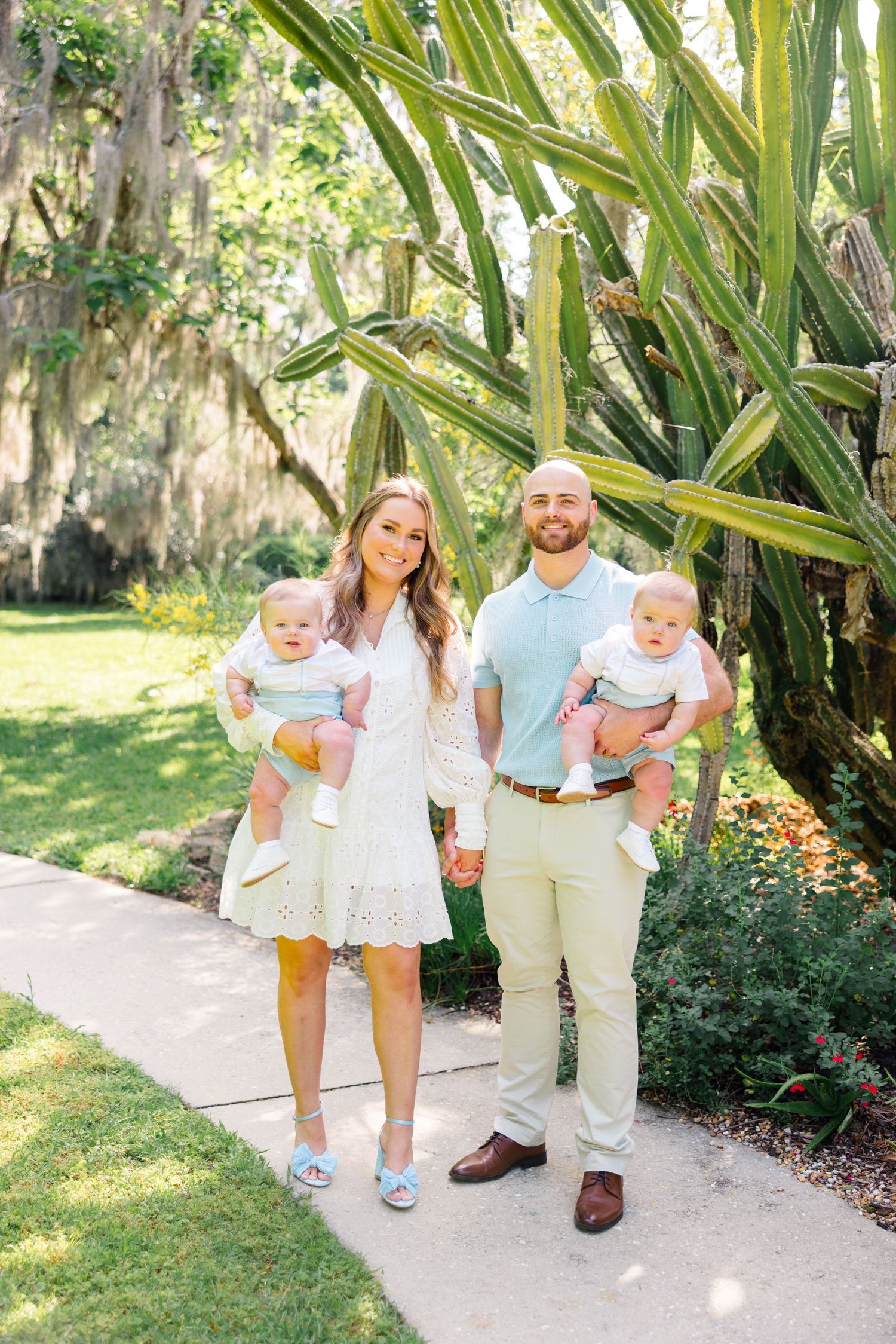 Family of three walking together on a garden path surrounded by lush tropical plants and greenery on a sunny day.
