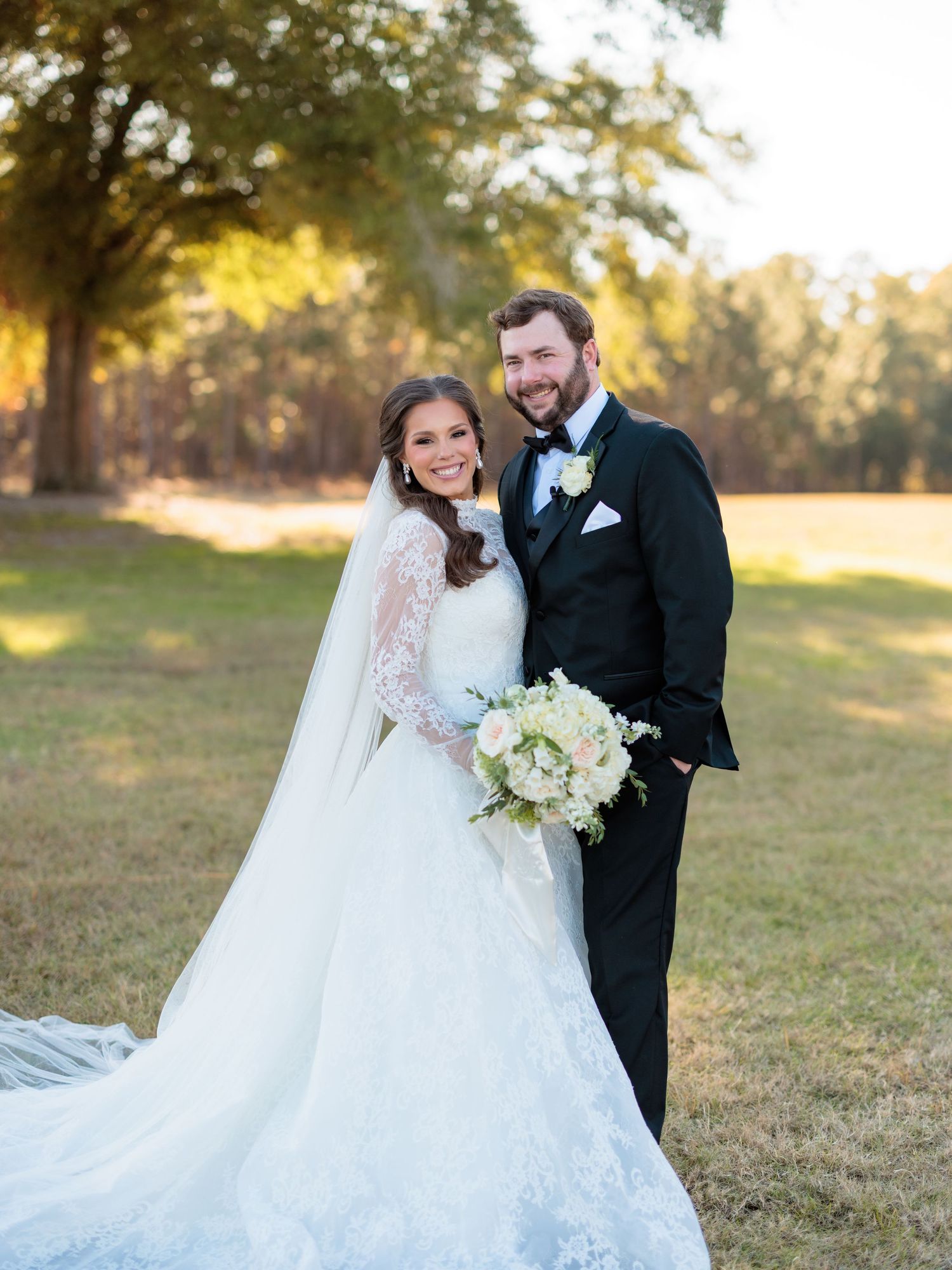 A wedding couple stands together outdoors in a natural setting with trees and sunlight, holding a white floral bouquet.