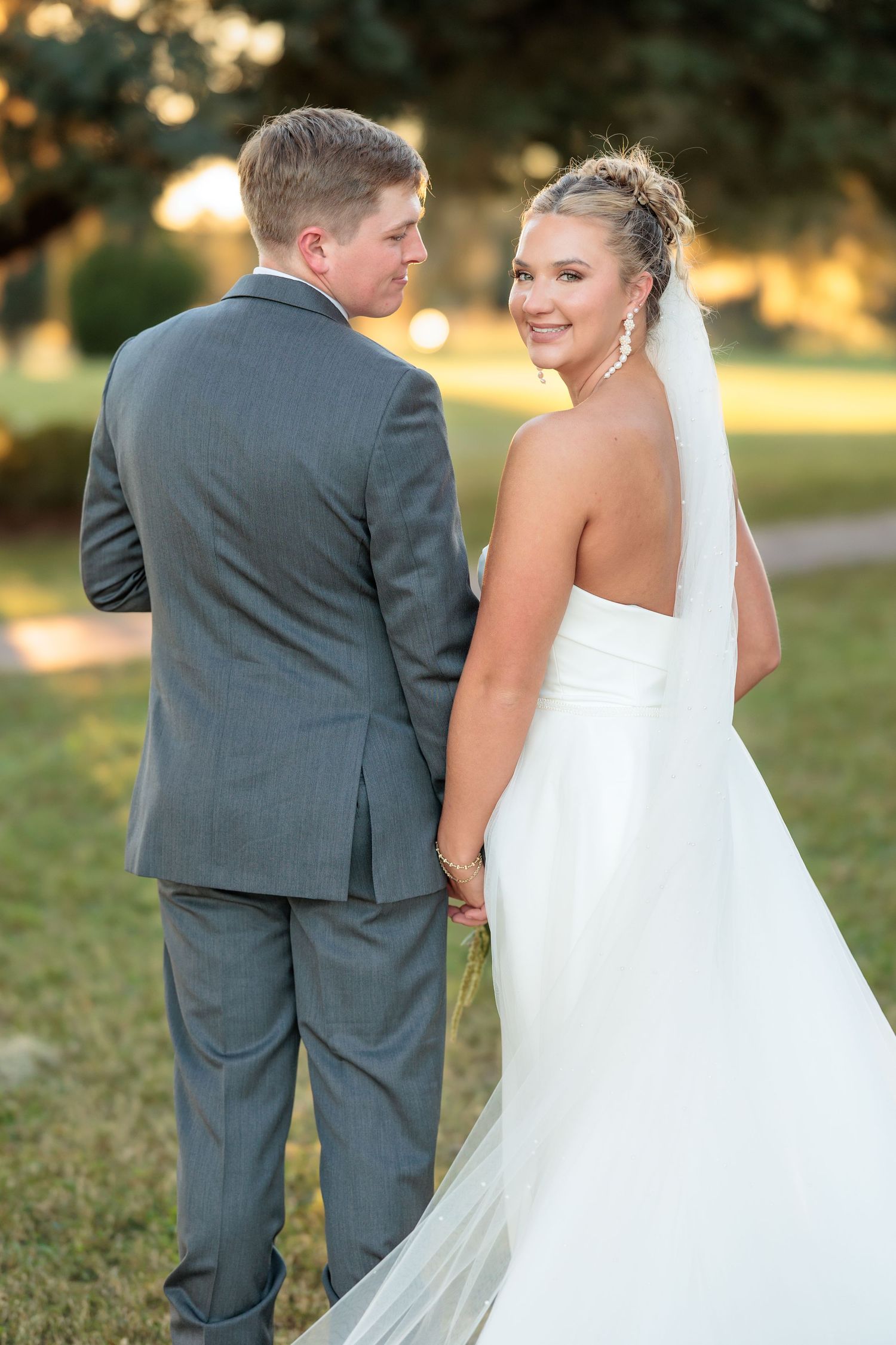 A bride in a white strapless gown and veil looks back at the camera while standing with her groom at sunset.