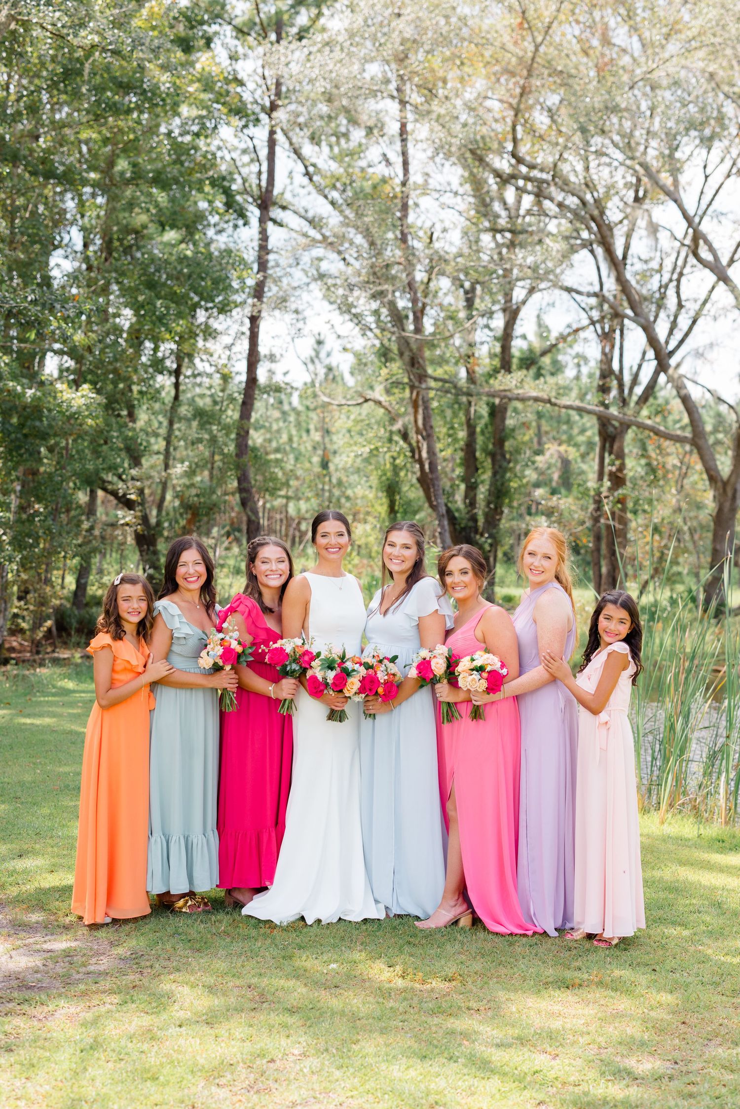 A bridal party poses outdoors in pastel dresses with colorful bouquets against a wooded backdrop.