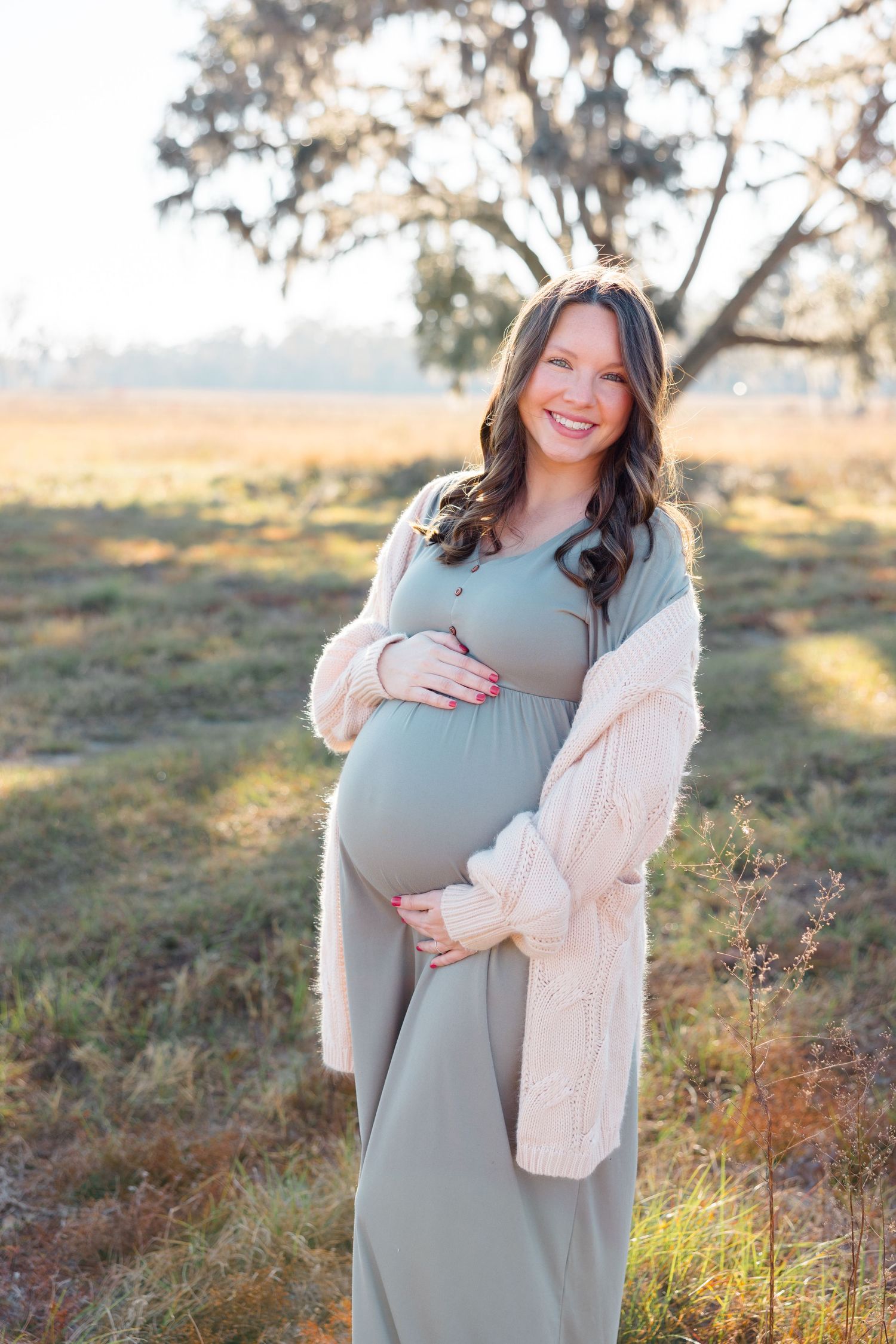 Expectant mother in a sage green dress poses outdoors wearing a cozy cream cardigan.