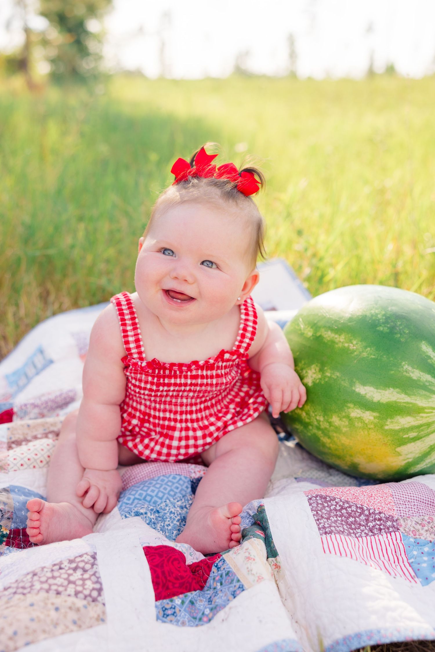 A baby in red gingham outfit smiles while sitting on a patchwork quilt next to a watermelon.