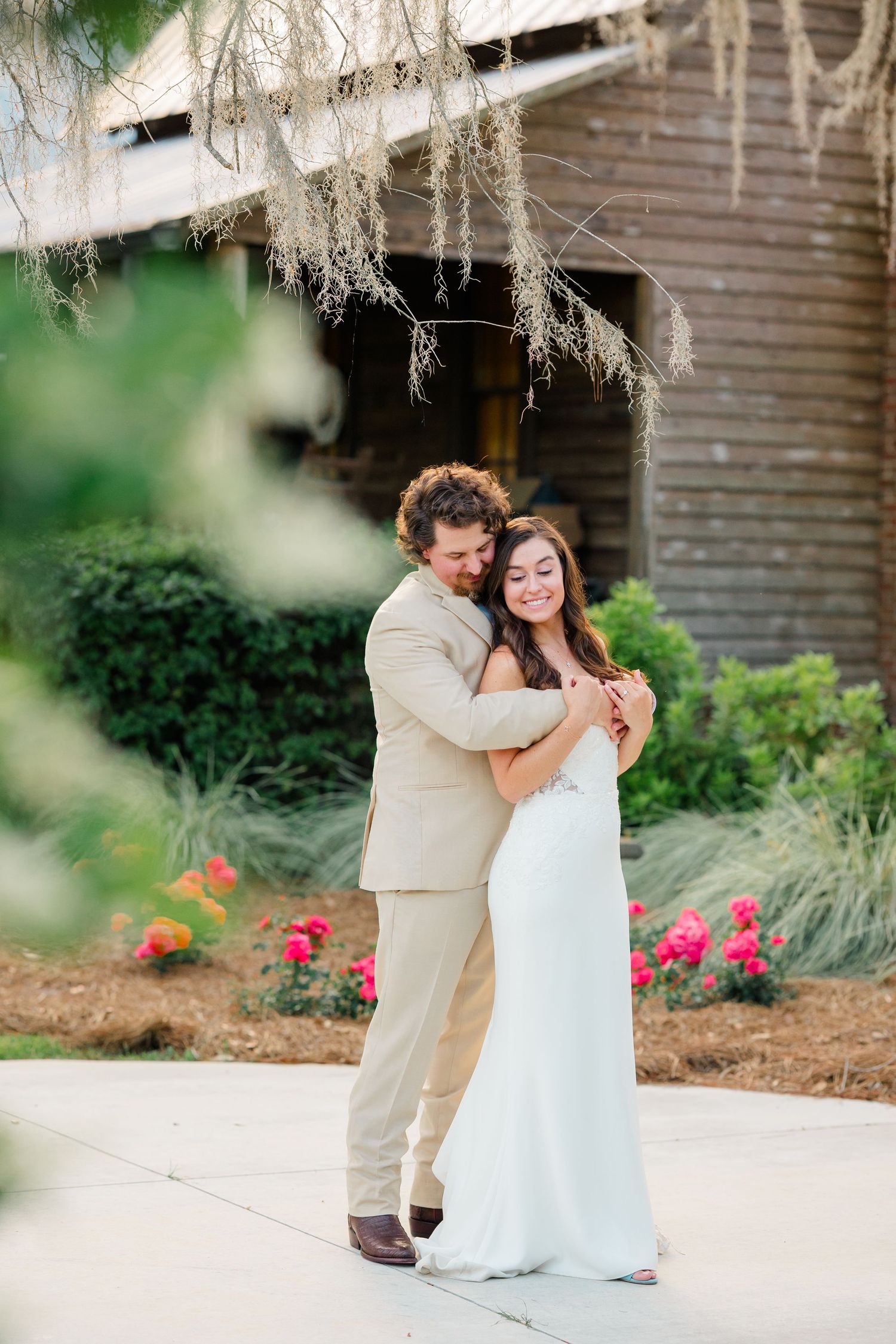 A couple in wedding attire shares a romantic moment under Spanish moss at an outdoor venue with pink flowers.