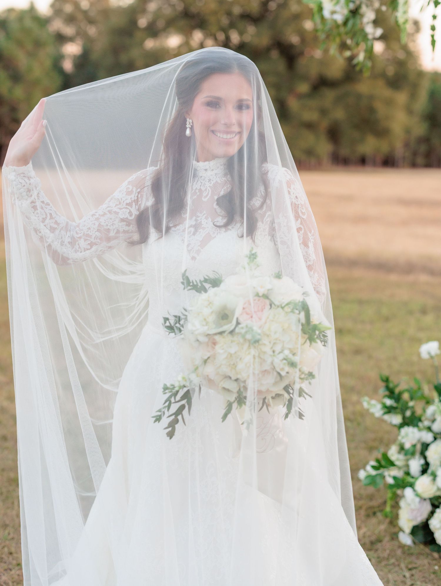 A bride wearing a delicate lace veil stands outdoors in natural lighting with soft floral accents.
