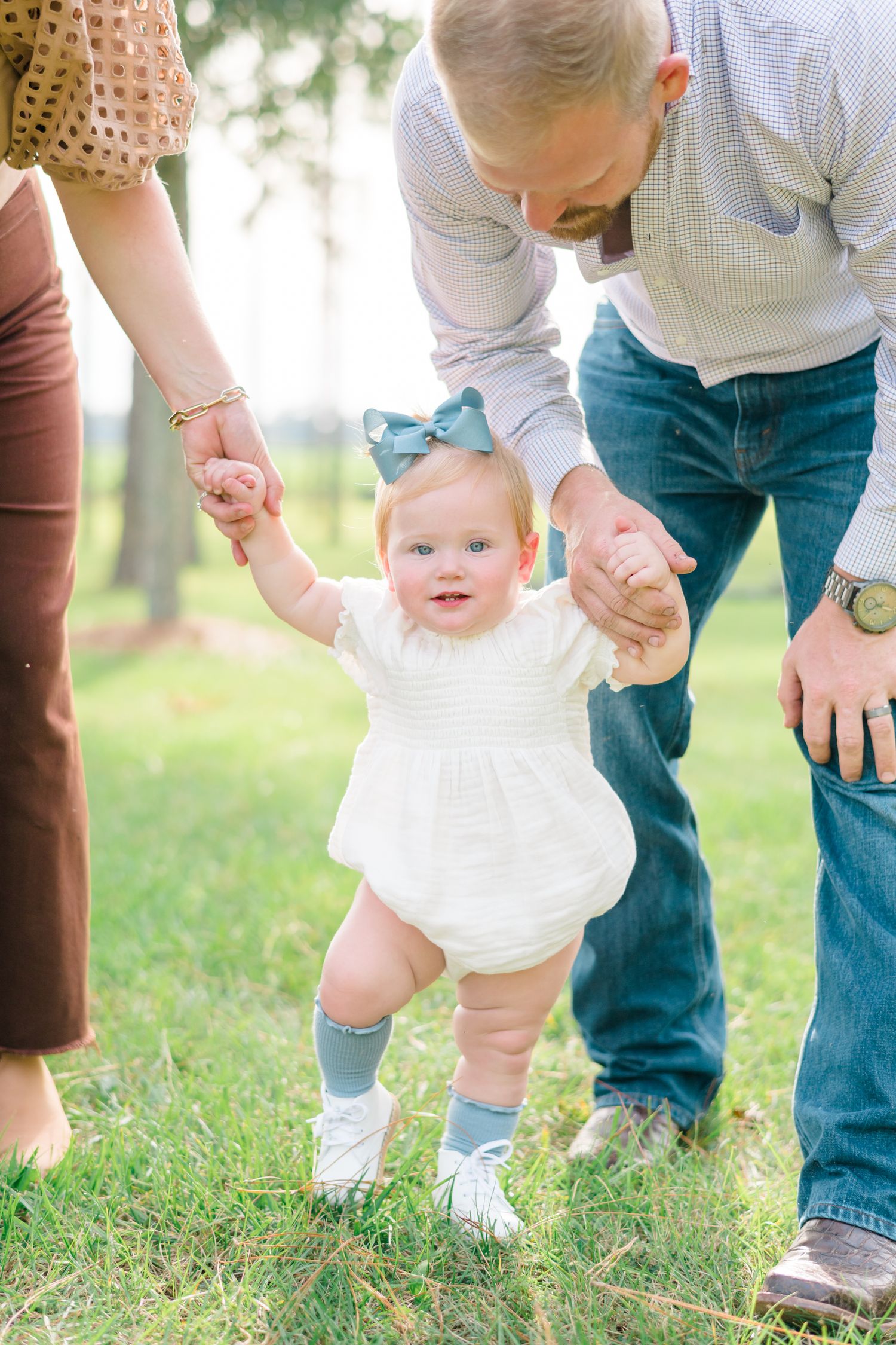 Series of walking steps of a baby in a white dress being supported by adults on grass.