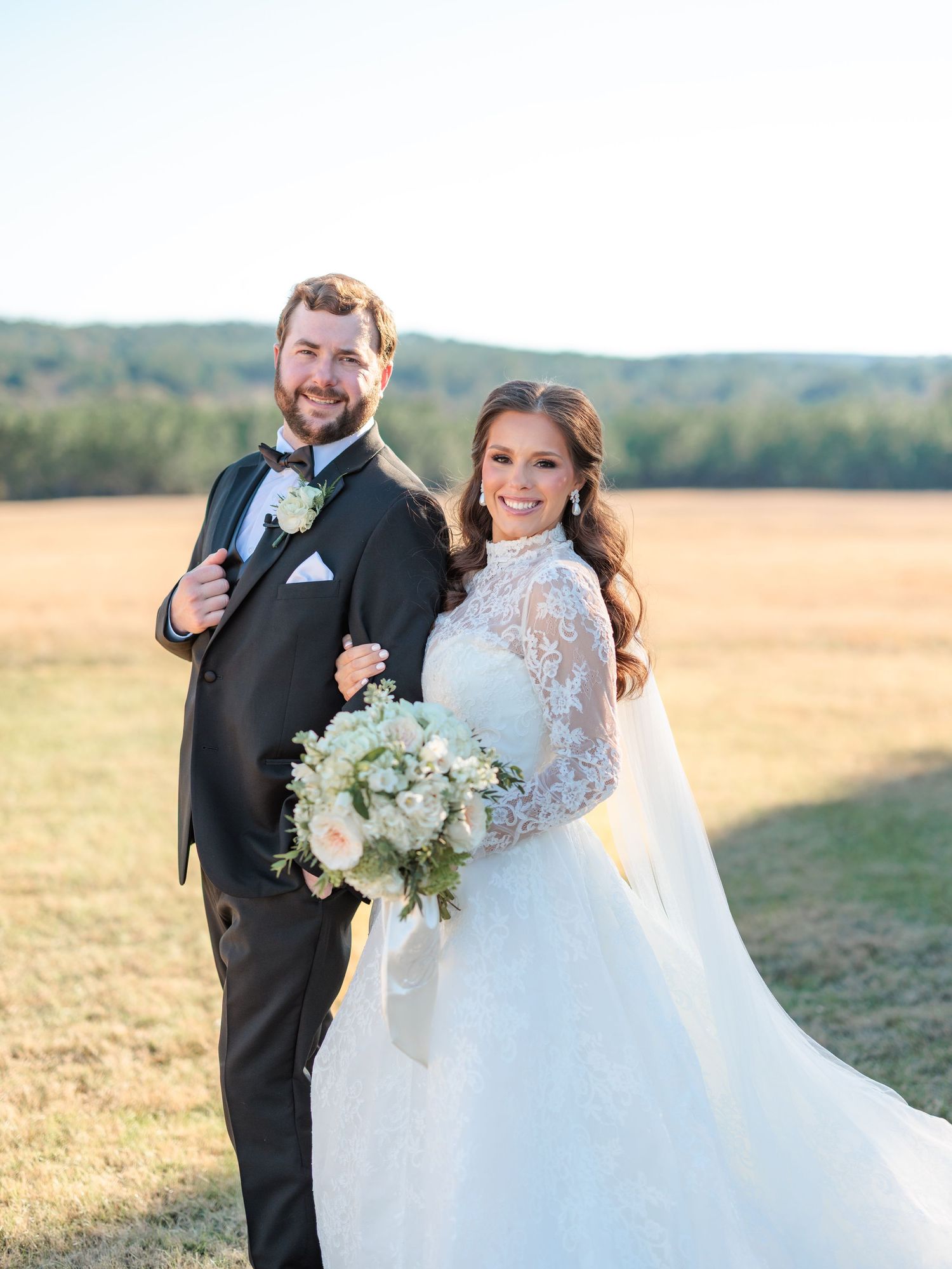 Newlywed couple poses together in a golden field during sunset while holding a white floral bouquet.