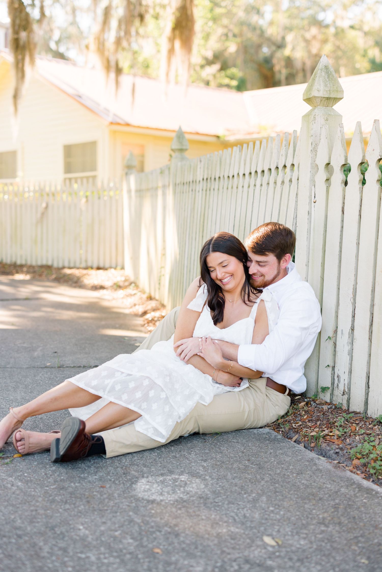 A couple relaxes on a sidewalk against a white picket fence in matching outfits.