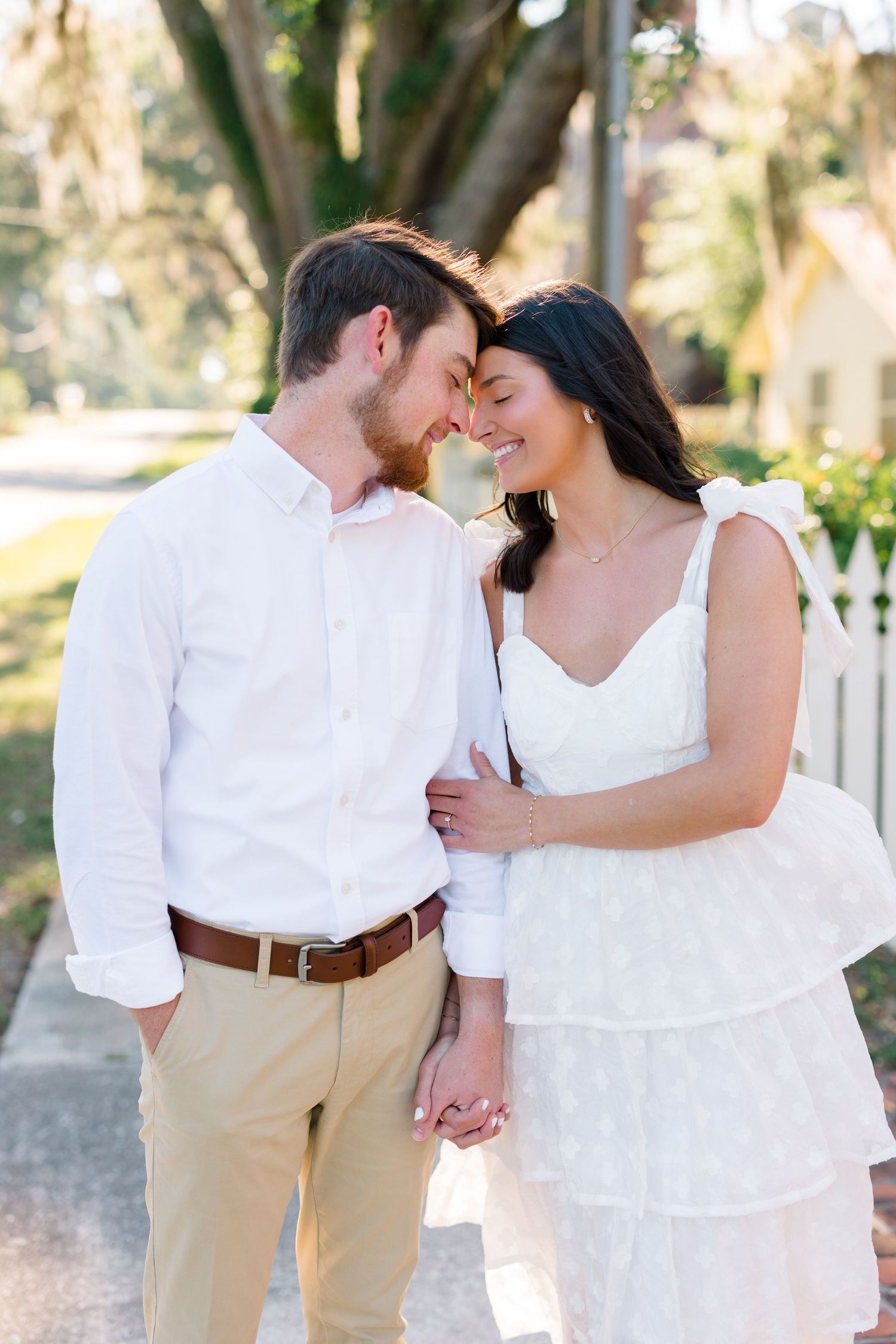 A romantic couple in white outfits share an intimate moment outdoors near a picket fence.