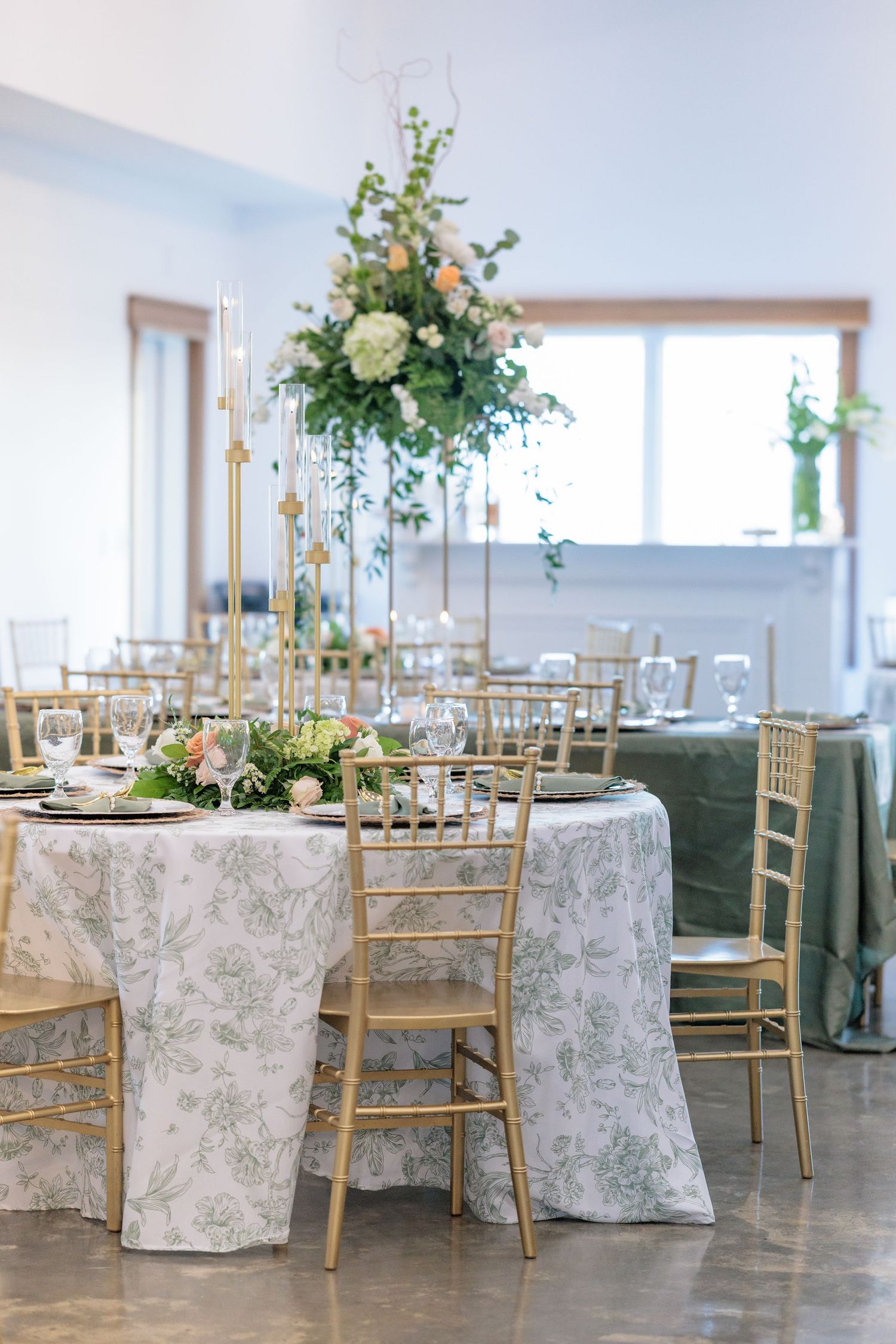 Elegant wedding reception setup with floral tablecloths, gold chiavari chairs and elevated floral centerpieces in bright space.