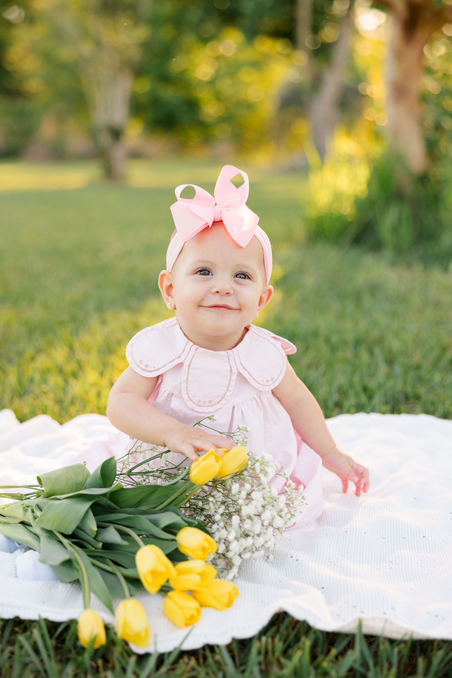 Toddler in pink dress and bow sits on white blanket with yellow tulips in outdoor park setting.