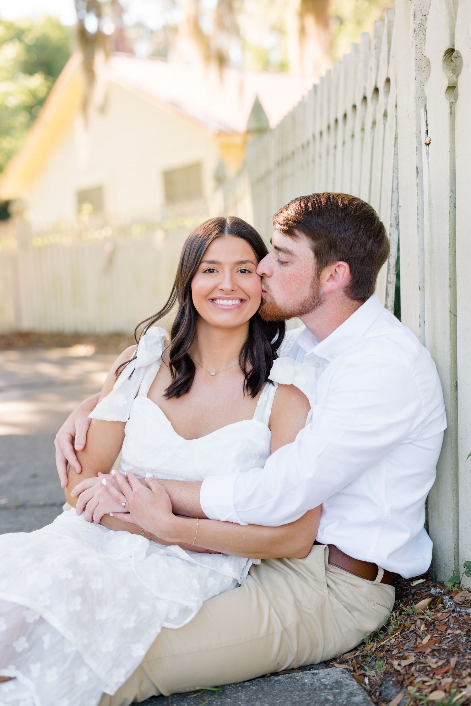 A couple sits on the ground by a white picket fence sharing an affectionate moment.