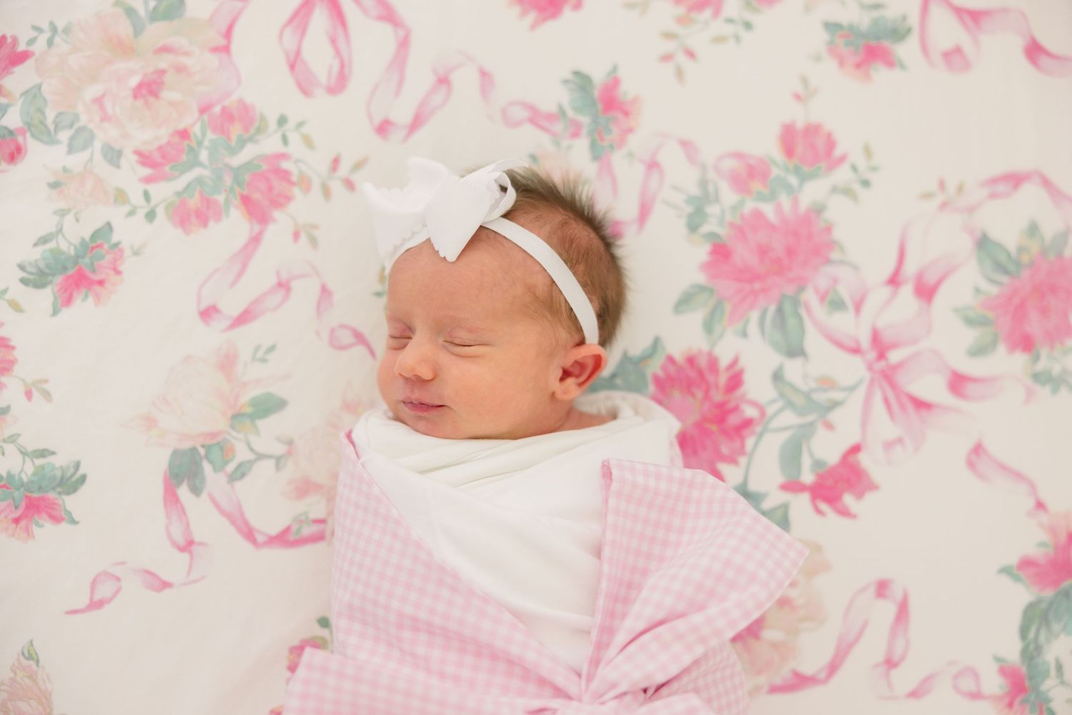 A newborn baby sleeps peacefully on pink floral bedding with a matching bow headband.