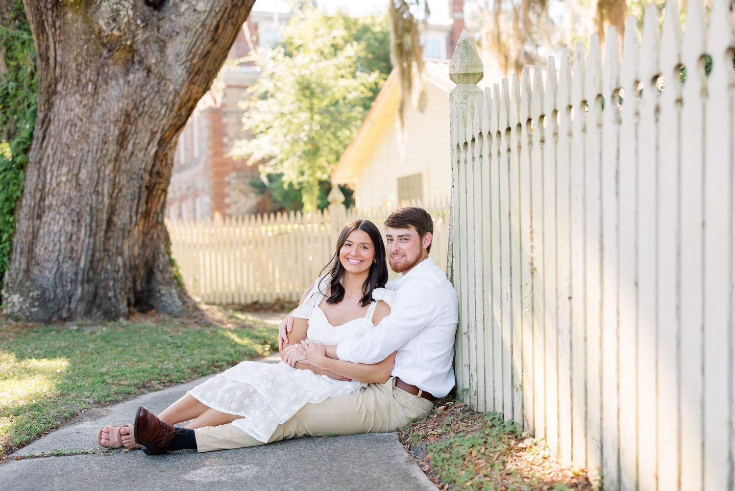 A couple sits together against a white picket fence under a large oak tree.