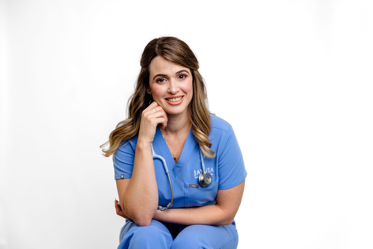 Healthcare professional in blue scrubs smiles while sitting and holding a stethoscope against white background.