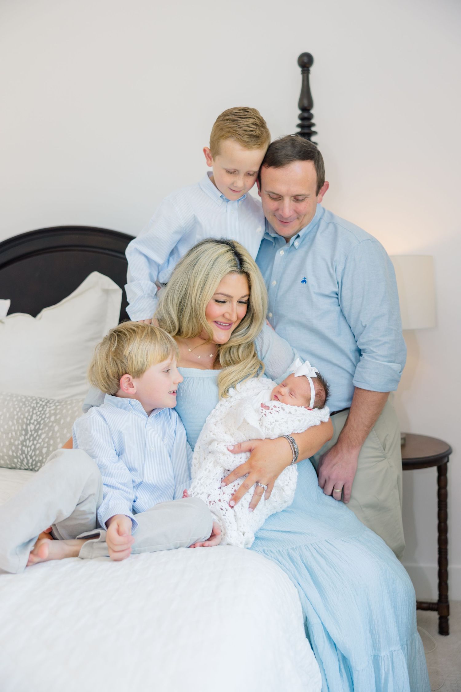 Family in soft blue clothing sitting together on white furniture sharing tender moments with a newborn baby.