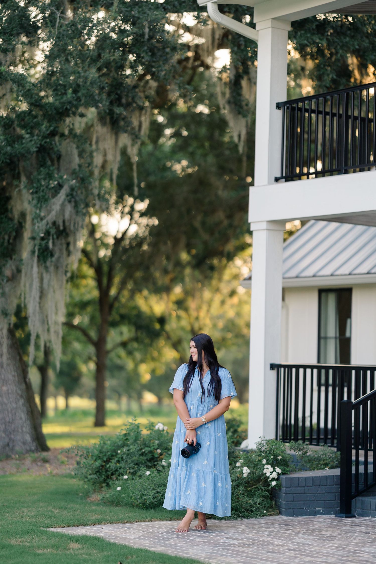 Woman in flowing blue dress stands on brick path near white building with Spanish moss draped trees in background.