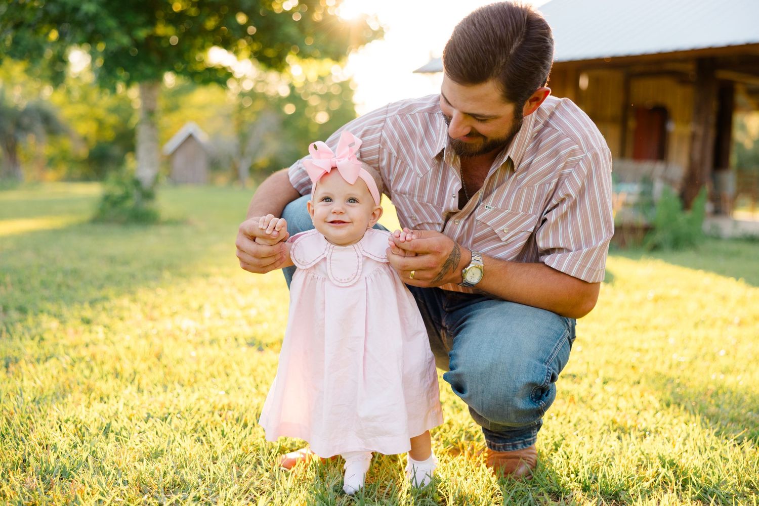 A parent and baby in pink dress share a tender outdoor moment at sunset during a family photoshoot.