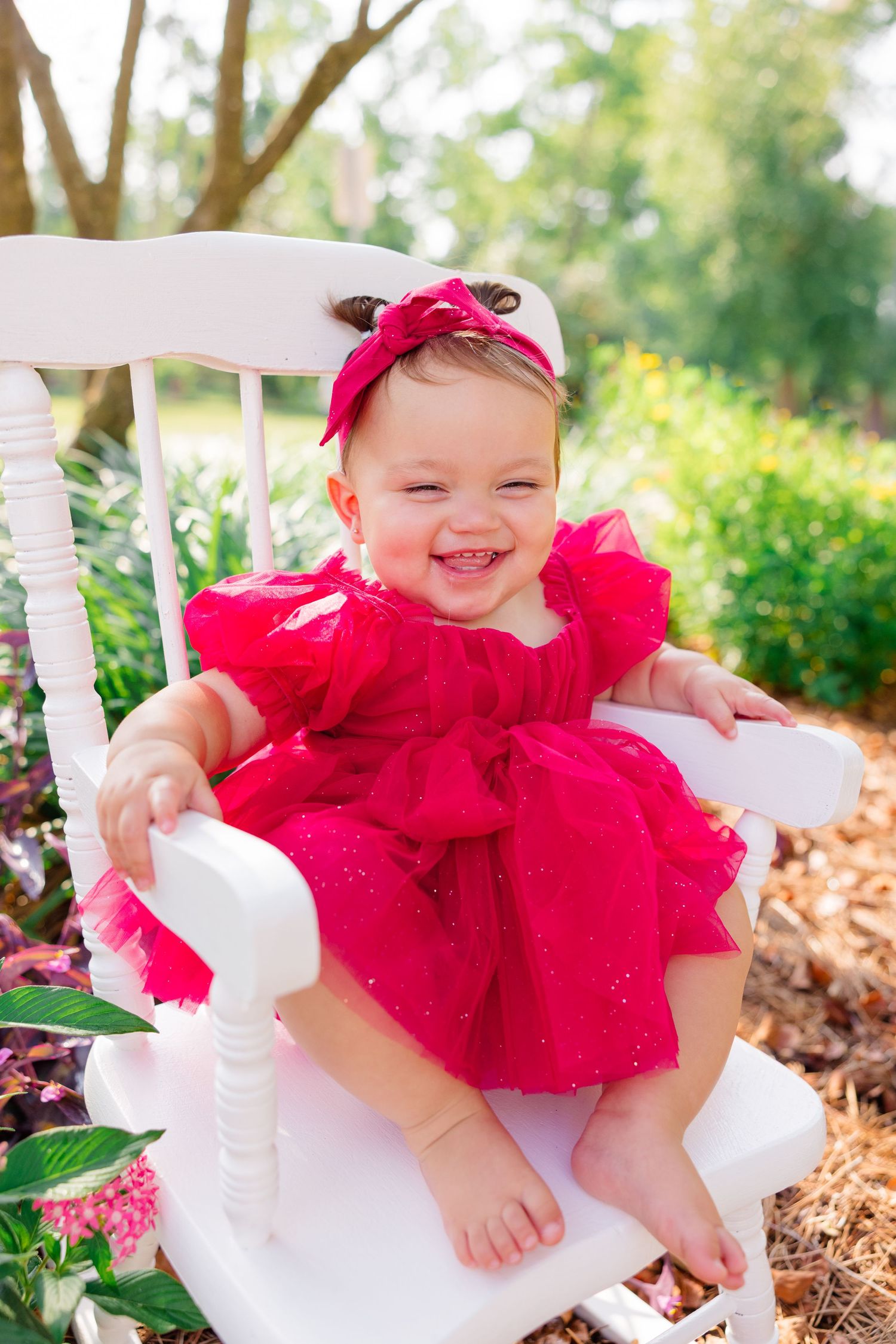 A smiling baby in a bright red tulle dress and bow sits on a white wooden chair outdoors.