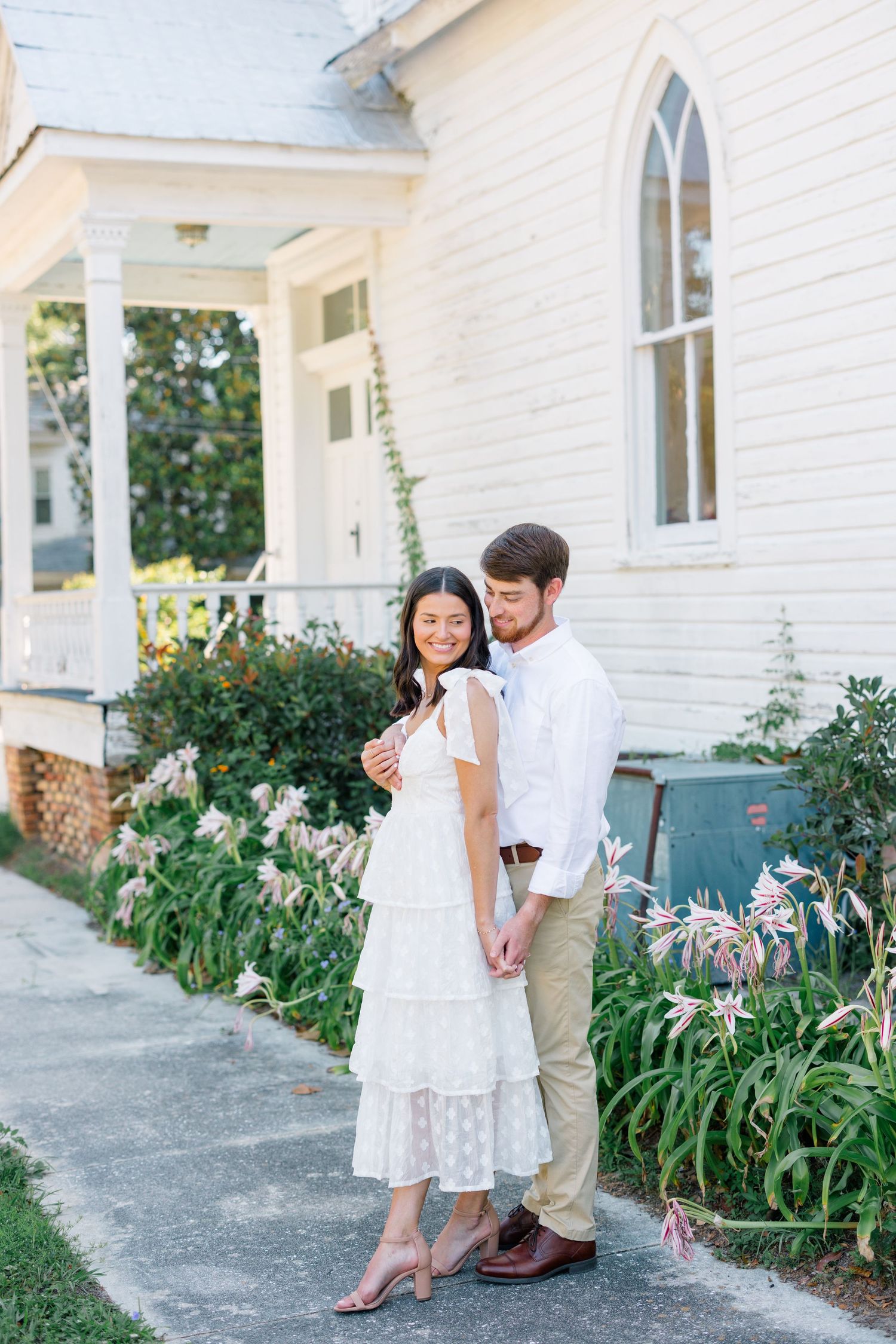 Two people in light colored outfits share a romantic moment against the white exterior of a church building.