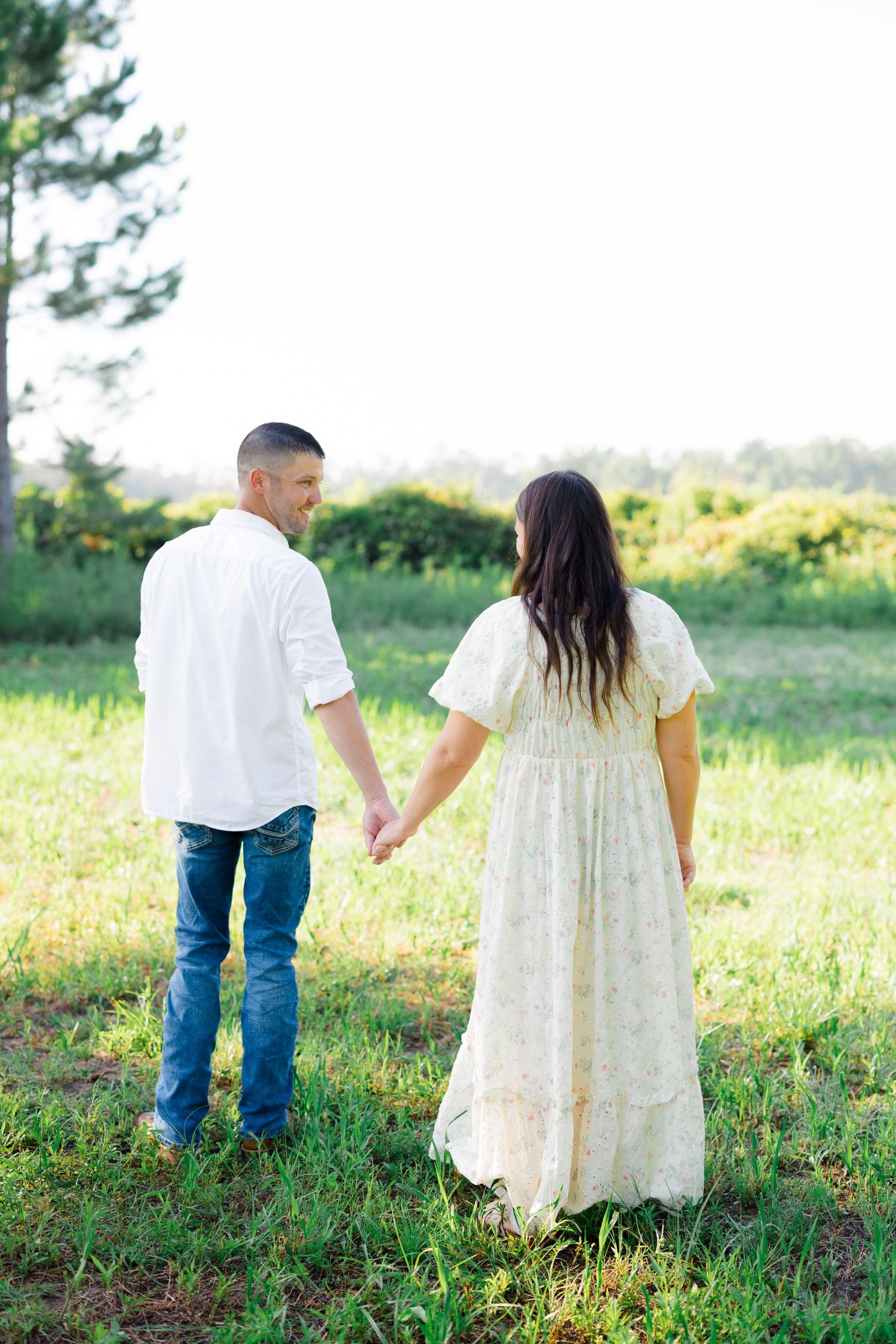 Couple walking hand in hand through a sunlit meadow wearing coordinated white outfits.